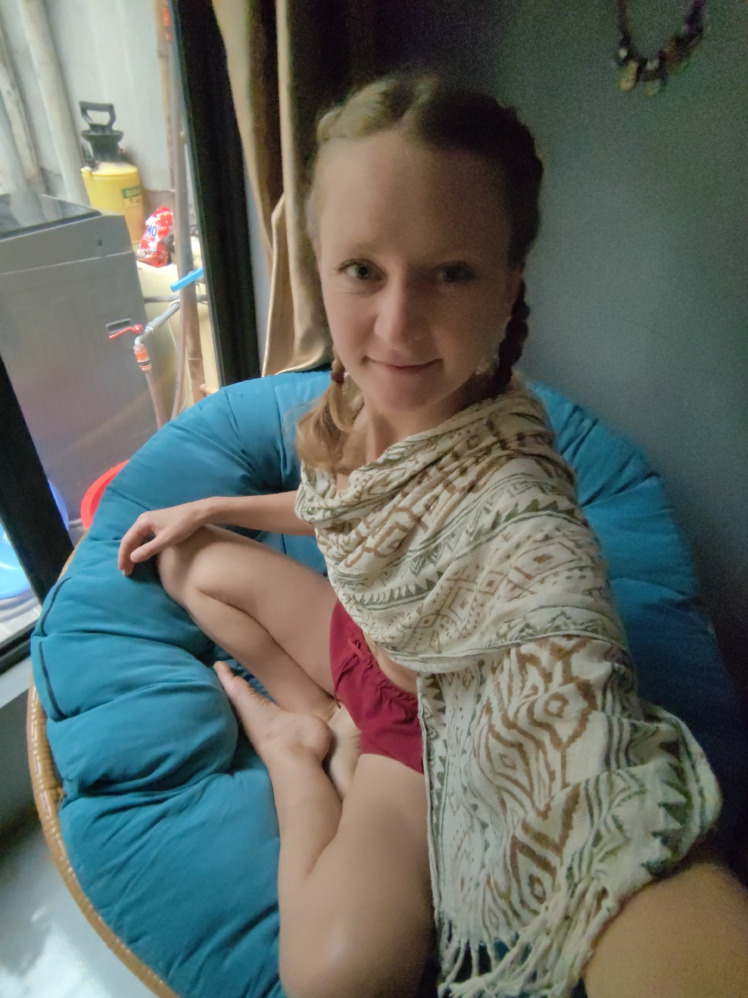 A young woman with light skin and braided hair sits relaxed in a round, blue cushioned chair with a rattan frame. She smiles gently, looking directly at the camera. She wears red shorts and a light-colored scarf with brown and green patterns, draped over her shoulders. Her bare feet are visible, resting in the chair.
The setting appears to be an indoor space. To the left, through what looks like a window or open area, a cluttered background is visible. This includes a grey utility box or appliance with a red handle and spigot, a yellow pump-action spray bottle, a red bag with the visible text "MO 10", and a clear blue tube. On the right, a brown curtain hangs, and above the woman's head, against a bluish-green wall, a decorative item resembling a necklace or garland is suspended. The lighting suggests it is daytime. The scene conveys a casual and relaxed atmosphere, likely in a home or personal dwelling in Hà Nội, Vietnam.