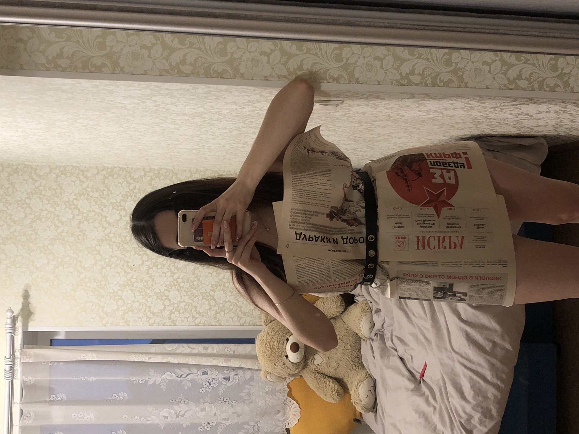 The image displays a person, likely a young woman, taking a mirror selfie in what appears to be a bedroom in Noviy, Russia. The individual's face is partially obscured by a light-colored smartphone held to capture the reflection. She is wearing a creatively constructed garment made from newspapers, resembling a dress or a top and skirt, cinched at the waist with a black belt featuring metal eyelets. Her long, dark hair cascades around her shoulders.
On the bed visible in the reflection, a light brown teddy bear with "I Love You" text on its foot rests on a yellow pillow and grey and white bedding. A small red object, possibly a pen or lipstick, is also on the bed. The background walls feature light-colored wallpaper with a subtle floral pattern. A white metal bed frame and a white patterned curtain are visible on the left side of the reflection. The indoor lighting suggests the photo was taken during the evening or night.
The newspaper garment displays various Cyrillic texts, indicating the use of Russian newspapers. Visible words and phrases, some mirrored or inverted, include "ПОБЕДА" (Victory), "ЗА" (For) alongside a red star emblem, "ЭКОЛОГИЯ" (Ecology), "НОВОСТИ" (News), "КУБЫ И КРАЙ" (Cubes and Edge/Region), "СТОИМОСТЬ" (Cost), and "ОТВЕТСТВЕННОСТЬ" (Responsibility). The scene captures a moment of personal style or playful experimentation.