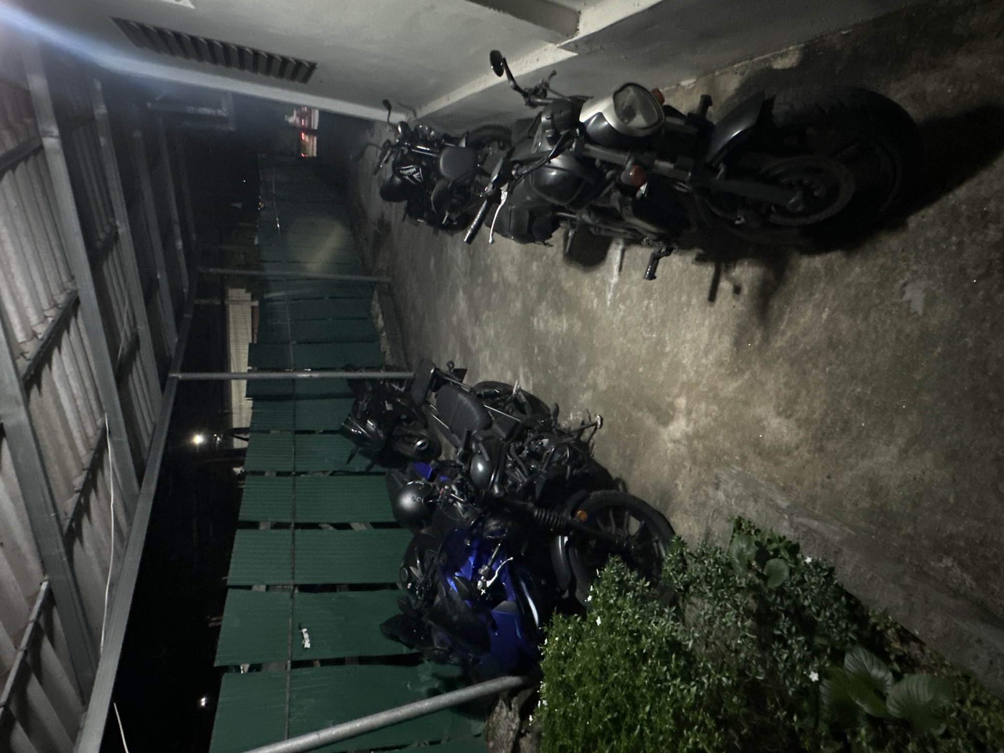 The image shows a row of parked motorcycles under a covered area, possibly a carport or an alleyway. The lighting is dim, suggesting it is nighttime or an overcast day. In the foreground, there is lush green foliage. The motorcycles are of various types and colors, with some appearing to be sportbikes and others cruiser-style bikes. One motorcycle in the foreground is black with a silver front fender and a round headlight. Another motorcycle to its left is blue and appears to be a sportbike, with a black helmet resting on its seat. Further down the row, more motorcycles are visible, some with luggage attached. The ground is paved with concrete, and there is a green corrugated metal fence in the background. The overall impression is that of a parking area for motorcycles in an urban setting, possibly Karon, Thailand, given the climate and vegetation.