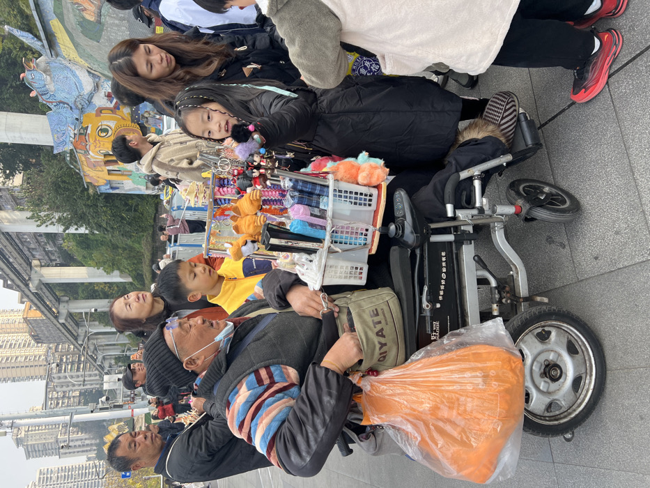 A man in a wheelchair, with a face mask, holds an orange plastic bag. A display of hair accessories and small stuffed animals is attached to the wheelchair. Around the man, several people stand, including a girl looking towards the camera. A woman with long brown hair stands behind the girl. In the background, there are buildings and a sky with white clouds.