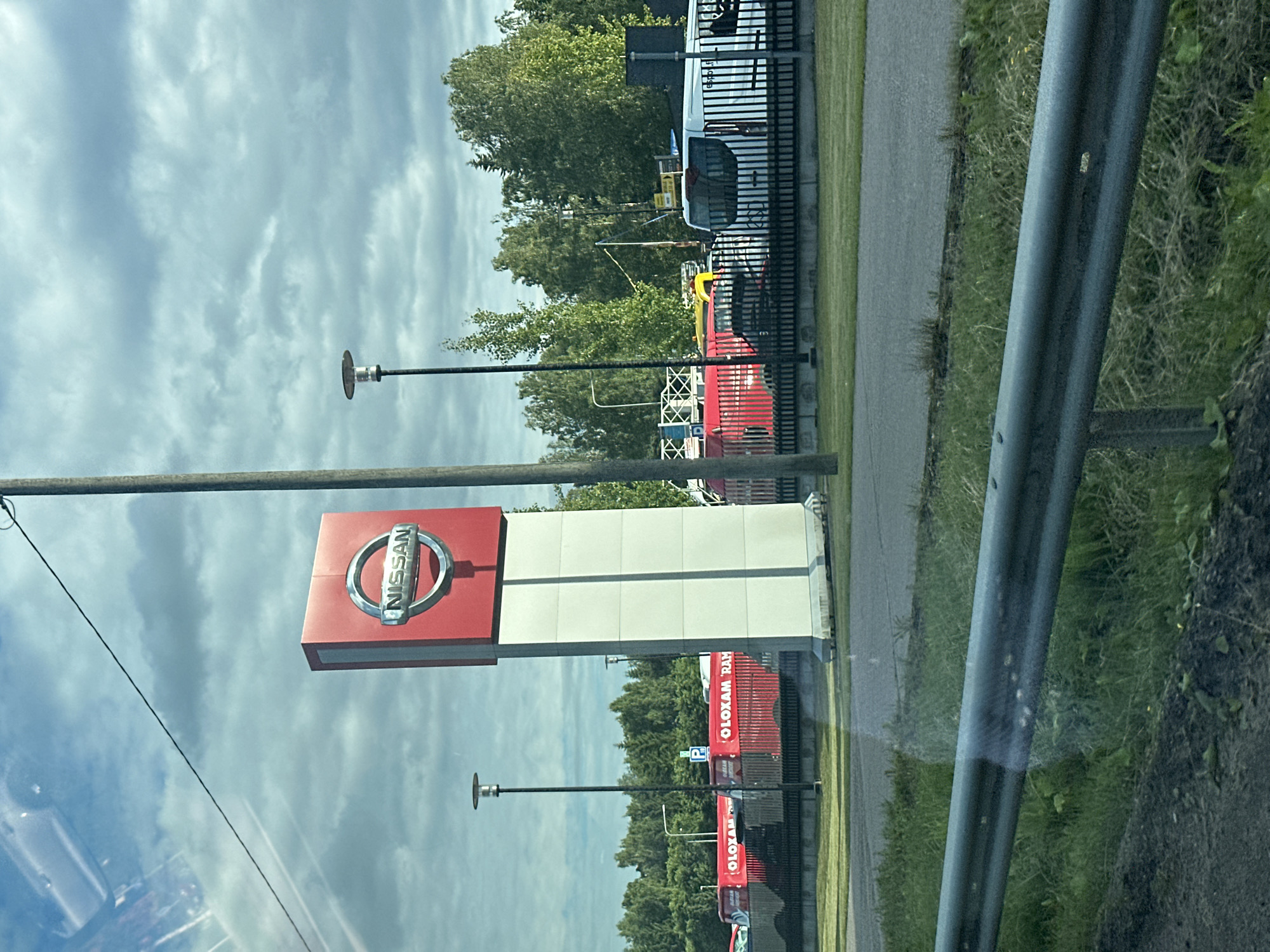 The image captures a Nissan dealership with a tall, red and white sign featuring the Nissan logo. In the background, several red vans with "OLOXAM RAM" printed on them are parked, along with other vehicles, including a white van and what appears to be a yellow truck. The dealership is set against a backdrop of lush green trees and a cloudy sky. A street lamp is visible near the Nissan sign, and a metal guardrail runs along the foreground, separating the road from a grassy area. The scene appears to be an outdoor commercial lot.