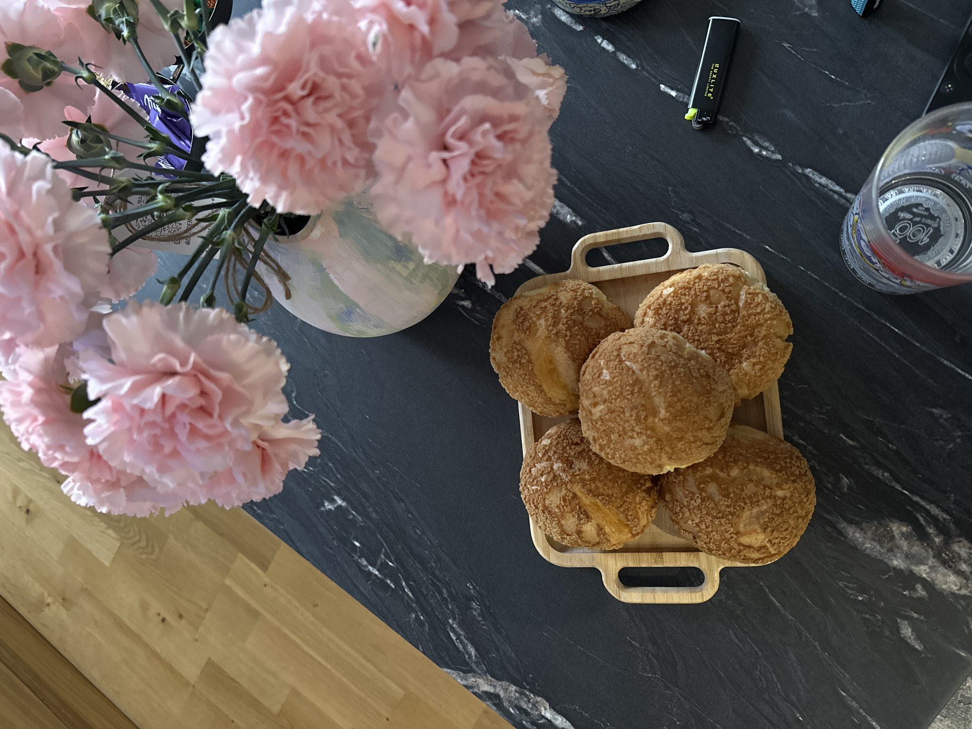 A top-down shot reveals a modern setting. Pink carnations in a light-colored, textured vase sit on a dark marble countertop that transitions to a wood floor. A light-colored bamboo tray holds five cream puffs. A black lighter and a drinking glass rest on the counter next to the tray. The scene is most likely located in Yerevan, Armenia, given the user's prompt.