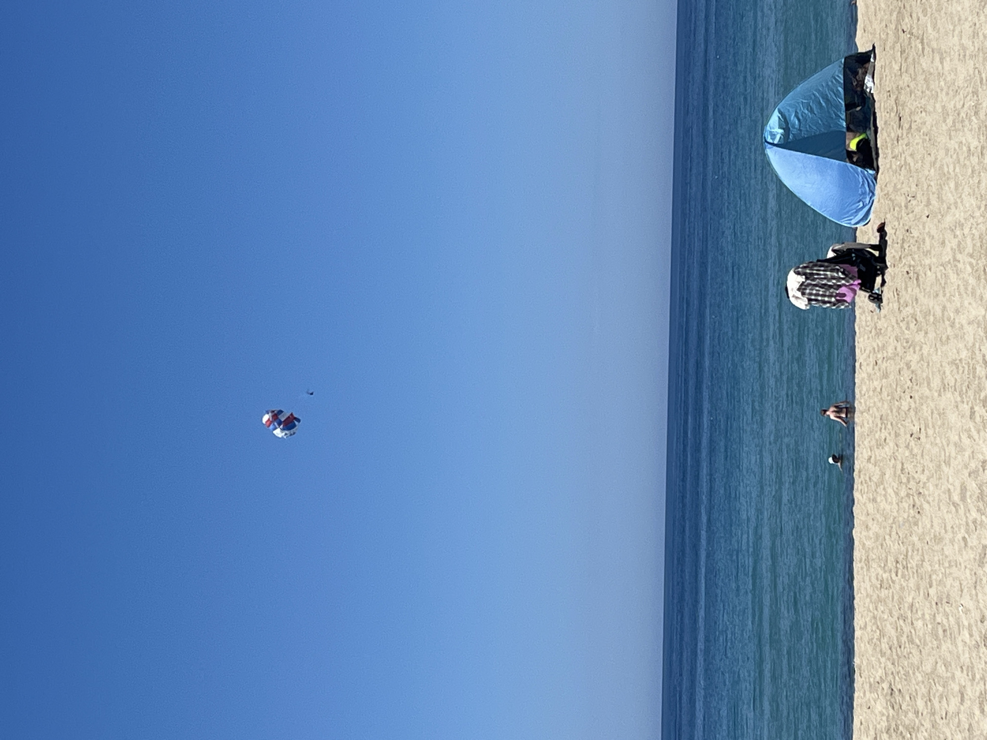 The image shows a view of a beach with the sea on the right and a clear blue sky dominating the left side of the frame. In the sky, there is a parasail with two people suspended beneath it. On the beach, there is a blue sun shelter, along with several people, some standing near the tent, and others in the water. The location is likely Alanya, Turkey, as it's a known destination for parasailing and beach tourism.