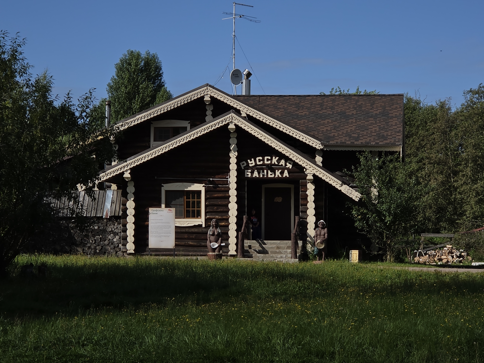 The image depicts a wooden bathhouse, identified by the Cyrillic inscription "РУССКАЯ БАНЬКА" above the entrance. The bathhouse has a distinctive roof with decorative trim and is surrounded by trees and a grassy area. Two human-like statues are positioned in front of the building: one to the left of the entrance appears to be a female figure, and another to the right is a male figure holding a basin. A sign with Russian text is visible to the left of the entrance. In the background, there is a stack of firewood and a fence. The sky is clear and blue. This location is the "Grafskie Bani" (Count's Baths) in the city of Peterhof, Russia.