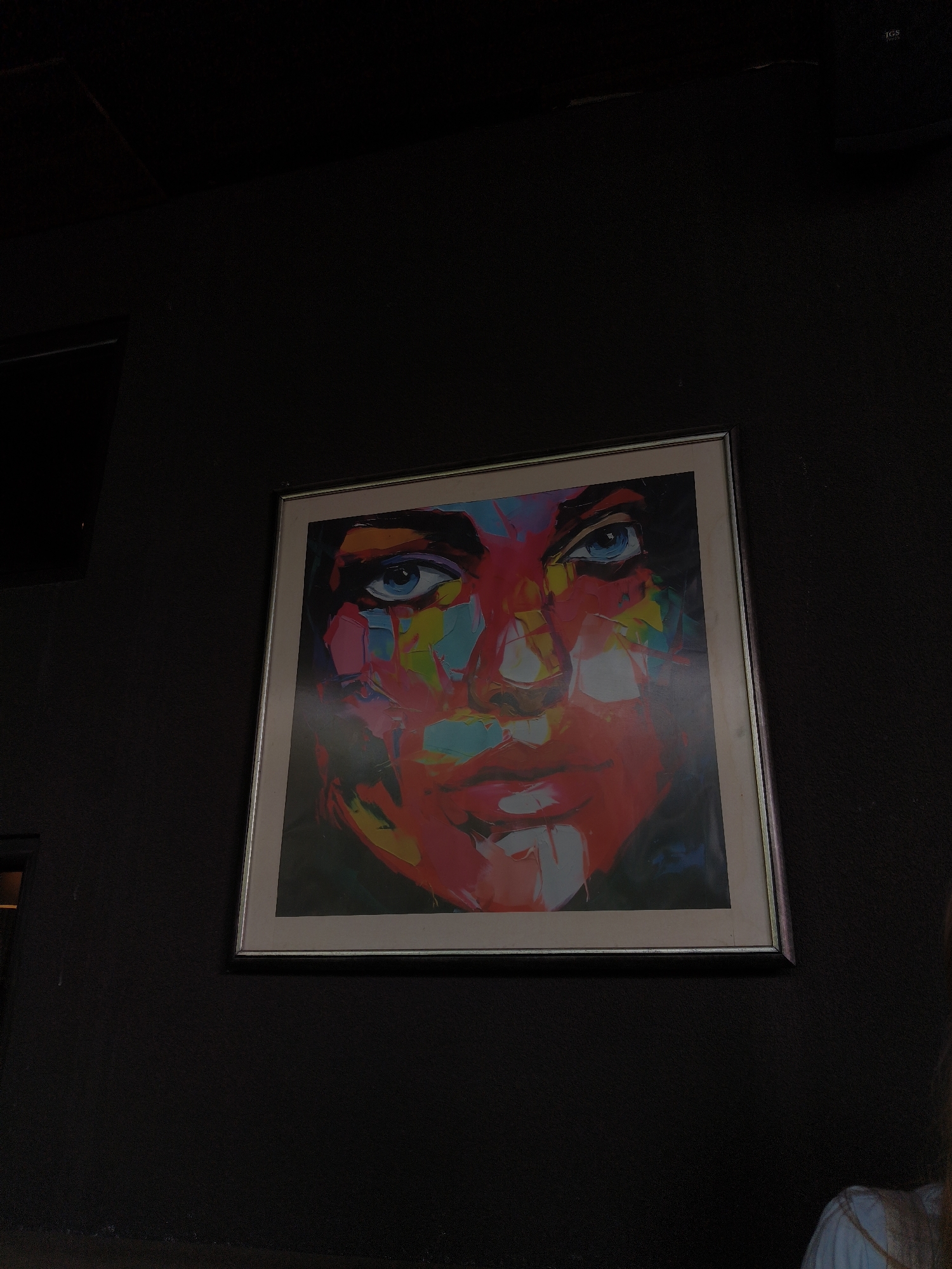 The image shows a framed abstract painting of a face hanging on a dark, textured wall. The painting is vibrant and uses bold strokes of color, including bright blues for the eyes, red for the face, and hints of yellow, green, and pink. The style is expressive and modern. The face appears to be looking directly at the viewer. The lighting is dim, suggesting an indoor setting, possibly a room or gallery. The framing of the painting is simple and silver-toned. In the bottom right corner, a small portion of a person is visible, wearing a light blue top with white trim. The overall mood is somewhat mysterious due to the dark background and the abstract nature of the artwork. No visible text is present in the image. The location context is Plovdiv, Bulgaria.