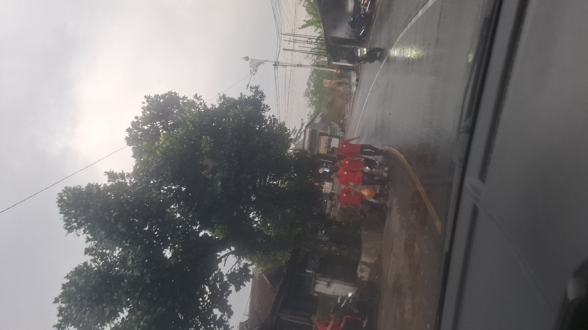 In Cilaku, Indonesia, on a cloudy day, a group of five young males wearing red shirts and dark pants walks along a wet asphalt road adjacent to a sidewalk lined with trees and modest buildings. One individual is carrying a light-colored object, possibly a balloon. A motorcycle is parked on the sidewalk to the left, and another motorcycle is seen riding on the road to the right. The scene appears to be captured from inside a vehicle, as evidenced by the dashboard visible in the foreground. The road is wet, suggesting recent or ongoing rain, with reflections visible on the surface.