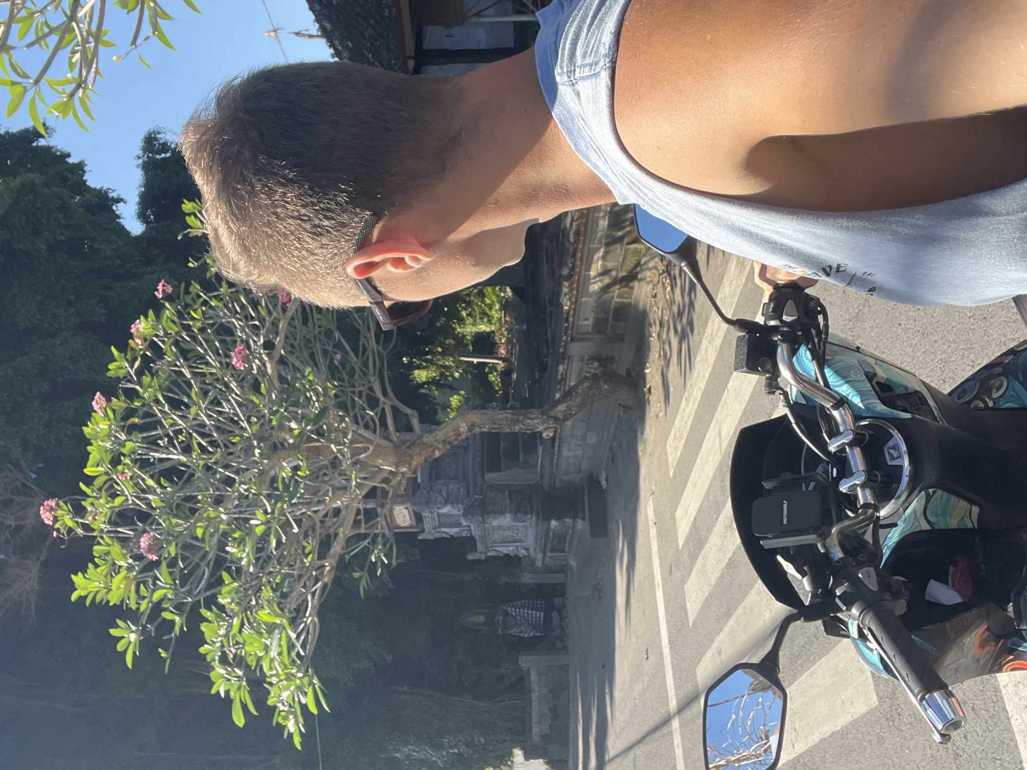 This media file shows a person riding a scooter in Sukawati, Indonesia. The person is viewed from behind, with their head turned to the side, wearing sunglasses and a light blue tank top. The scooter has a colorful, abstract pattern on its body and a black phone mount on the handlebars. In the background, there is a tree with pink flowers, lush green foliage, and traditional Indonesian architecture, including a stone structure that appears to be part of a temple or shrine. The scene is set on a paved road with a visible pedestrian crossing. The lighting suggests it is a bright, sunny day, likely in the morning or afternoon, with clear blue skies. The overall impression is one of a casual ride through a scenic, culturally rich environment.