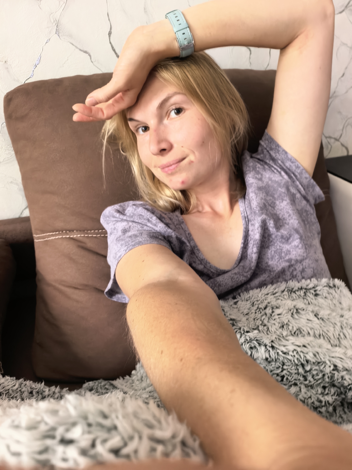 A fair-skinned woman with blonde hair is lying on a brown, upholstered couch covered with a gray and white fluffy blanket. She wears a lavender-colored t-shirt and a light blue wristwatch. Her right arm is raised, with her hand on her forehead. She is looking at the camera with a slight smile. The background features a light-colored wall with a marble-like pattern. The location is likely indoors, but there is no other information to determine if it is in the city of Sterlitamak, Russia.