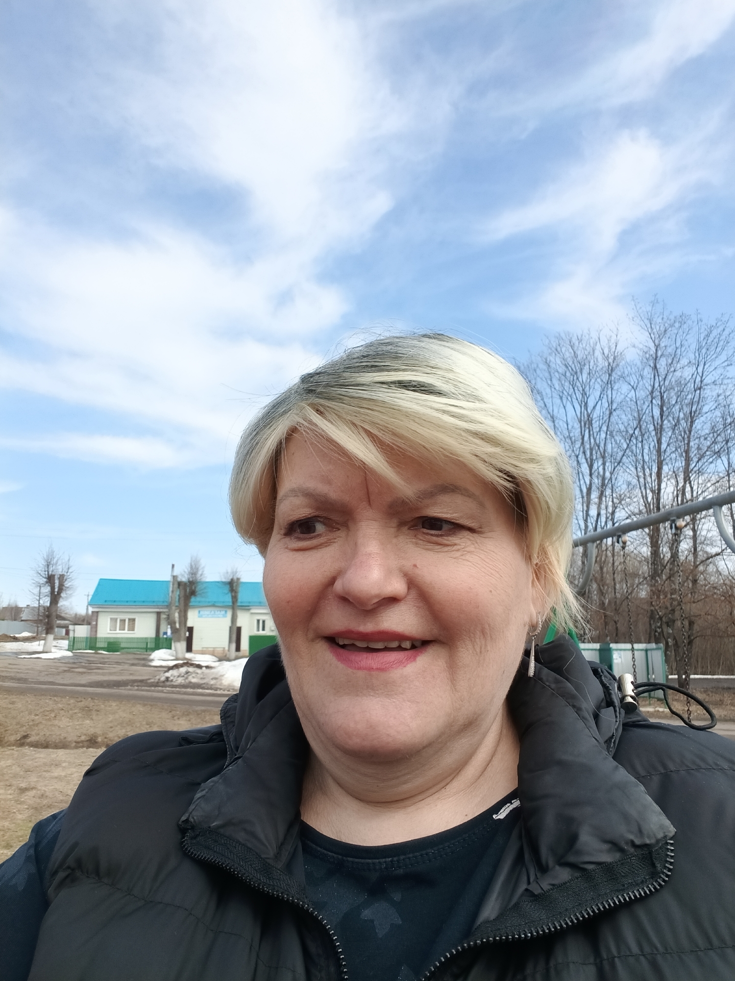 A middle-aged woman with short, blonde hair is smiling at the camera. She is wearing a black puffer vest and earrings. Behind her is a view of the Russian city Красный Маяк in what appears to be early spring. A light-colored building with a blue roof stands in the background, along with bare trees and a playground swing set. The sky is blue with scattered clouds. The ground shows patches of grass and some remnants of snow.