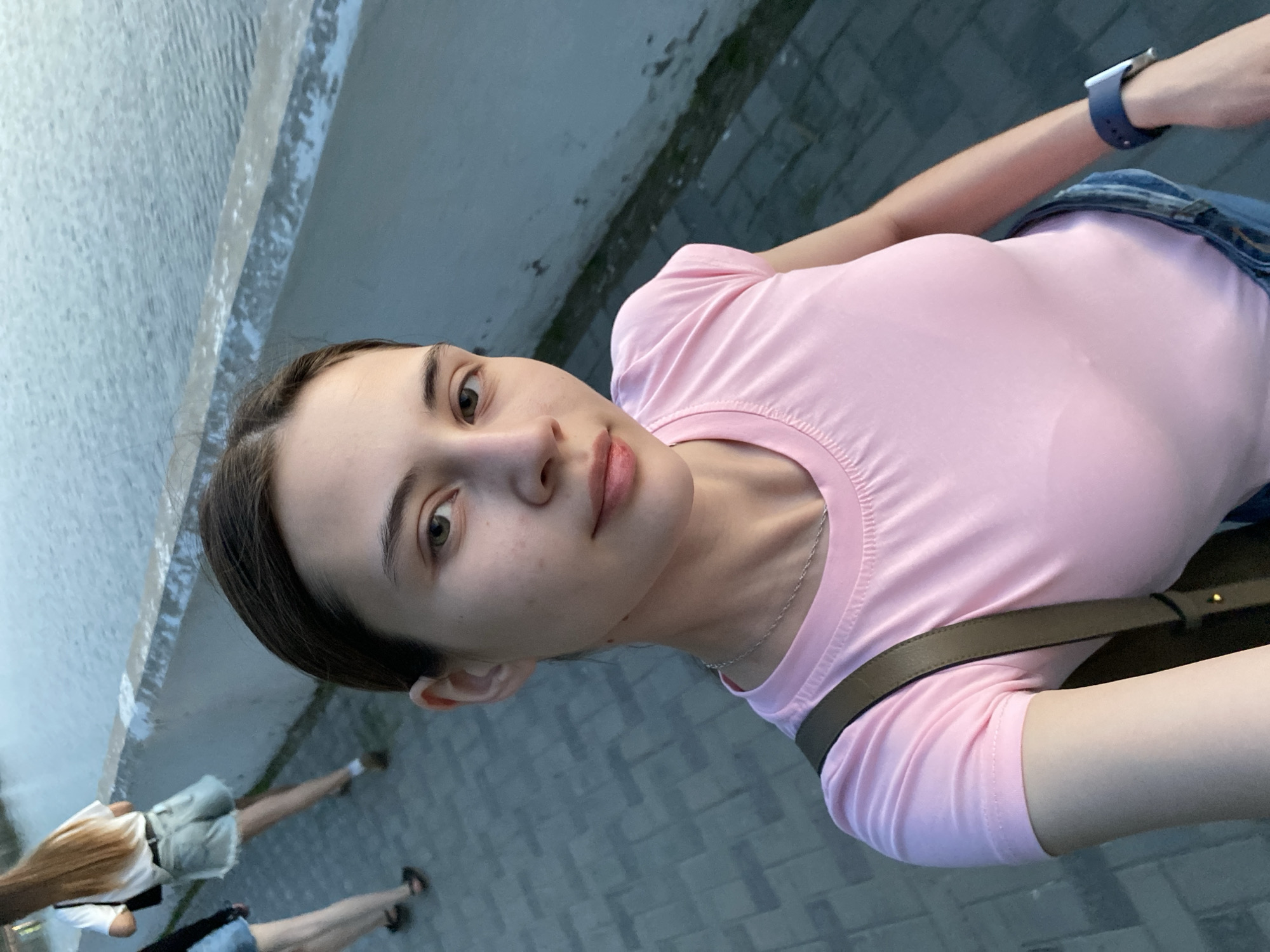 A young woman with brown hair pulled back poses for a selfie outdoors. She is wearing a pink t-shirt, jeans, and a blue smart watch on her left wrist. A dark-colored shoulder bag is slung across her body. She has fair skin, brown eyes, and a neutral expression.
The background includes a light grey stone wall or embankment, and a grey brick-paved walkway where other people are walking. The people visible in the lower left corner seem to be wearing summer clothing. The presence of the embankment suggests this location might be near water.
The location appears to be in a city, based on the paved walkway and other people present, and there is a possibility that the location is Votkinsk, Russia.