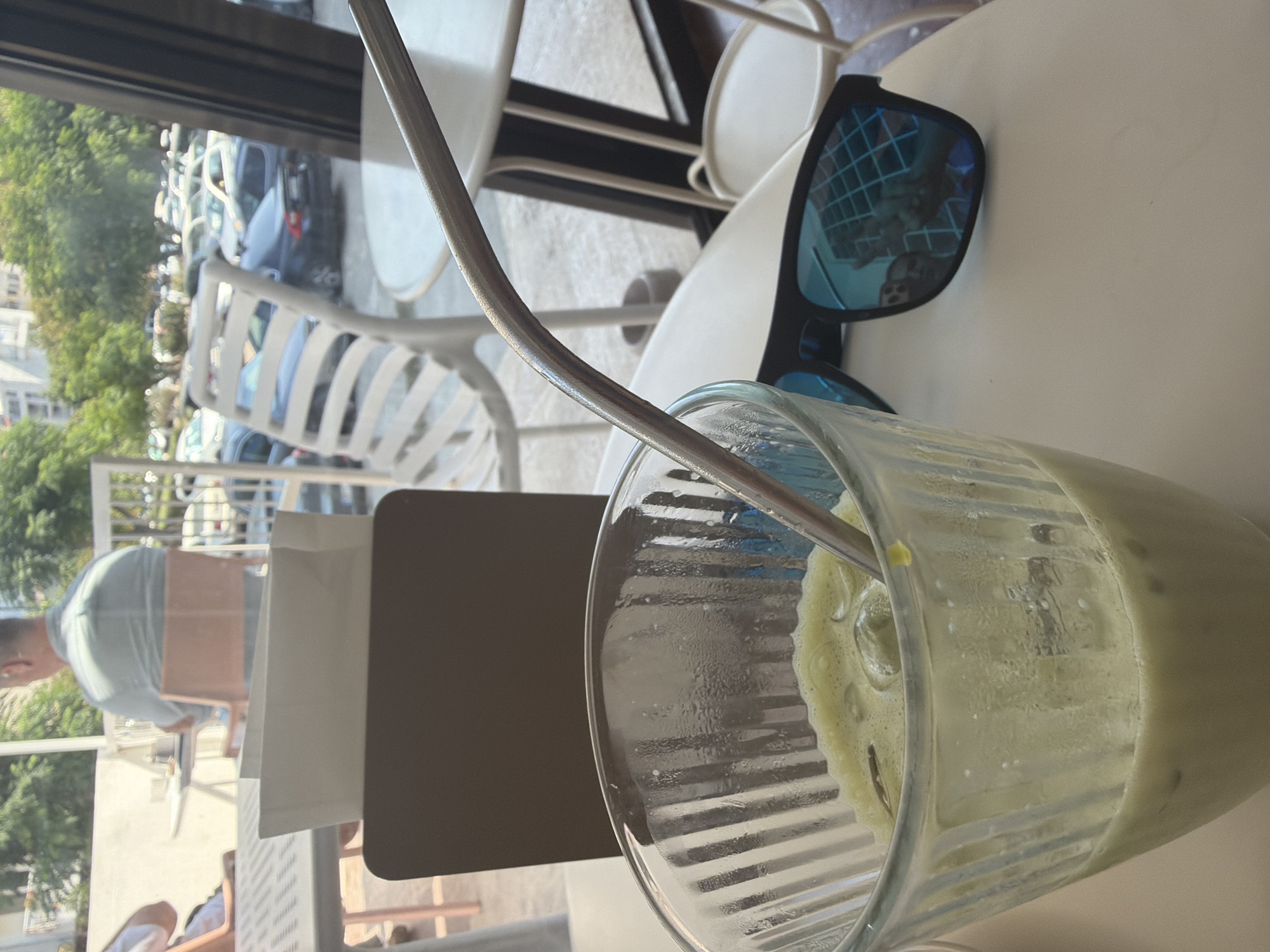 The image depicts a close-up view of a table setting at an outdoor cafe. In the foreground, a glass filled with a pale green, frothy beverage, likely a matcha latte, rests on a white table. Ice cubes are visible within the drink, and a metallic straw is inserted. To the right of the glass, a pair of black sunglasses with blue mirrored lenses is placed on the table, reflecting a grid-like structure, possibly a ceiling or outdoor awning.
In the background, through what appears to be a glass window or opening, a street scene unfolds. Cars are parked along a street lined with trees, and several outdoor chairs and tables are visible, some occupied by people. The overall ambiance suggests a relaxed daytime setting in a city.
While the specific location of Bar, Montenegro cannot be confirmed solely from the visual elements of the image, the presence of outdoor cafes, parked cars, and lush greenery are consistent with urban environments in coastal Mediterranean regions.