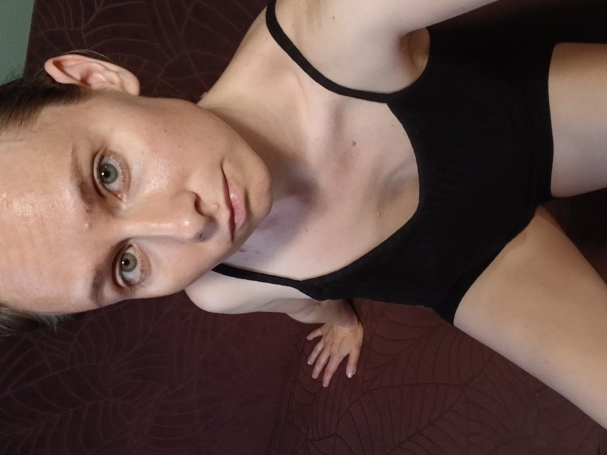 A young Caucasian woman with light skin, green eyes, and light brown hair pulled back is shown from the chest up, angled slightly towards the viewer. She is wearing a black tank top with thin straps. The woman is sitting on a dark brown, textured couch. Her left hand is resting on the couch behind her. The background consists of the couch and a dark wall. The lighting is soft, highlighting her face and upper body. The location is an indoor residential setting. There is no indication of the specific location within Yekaterinburg, Russia.