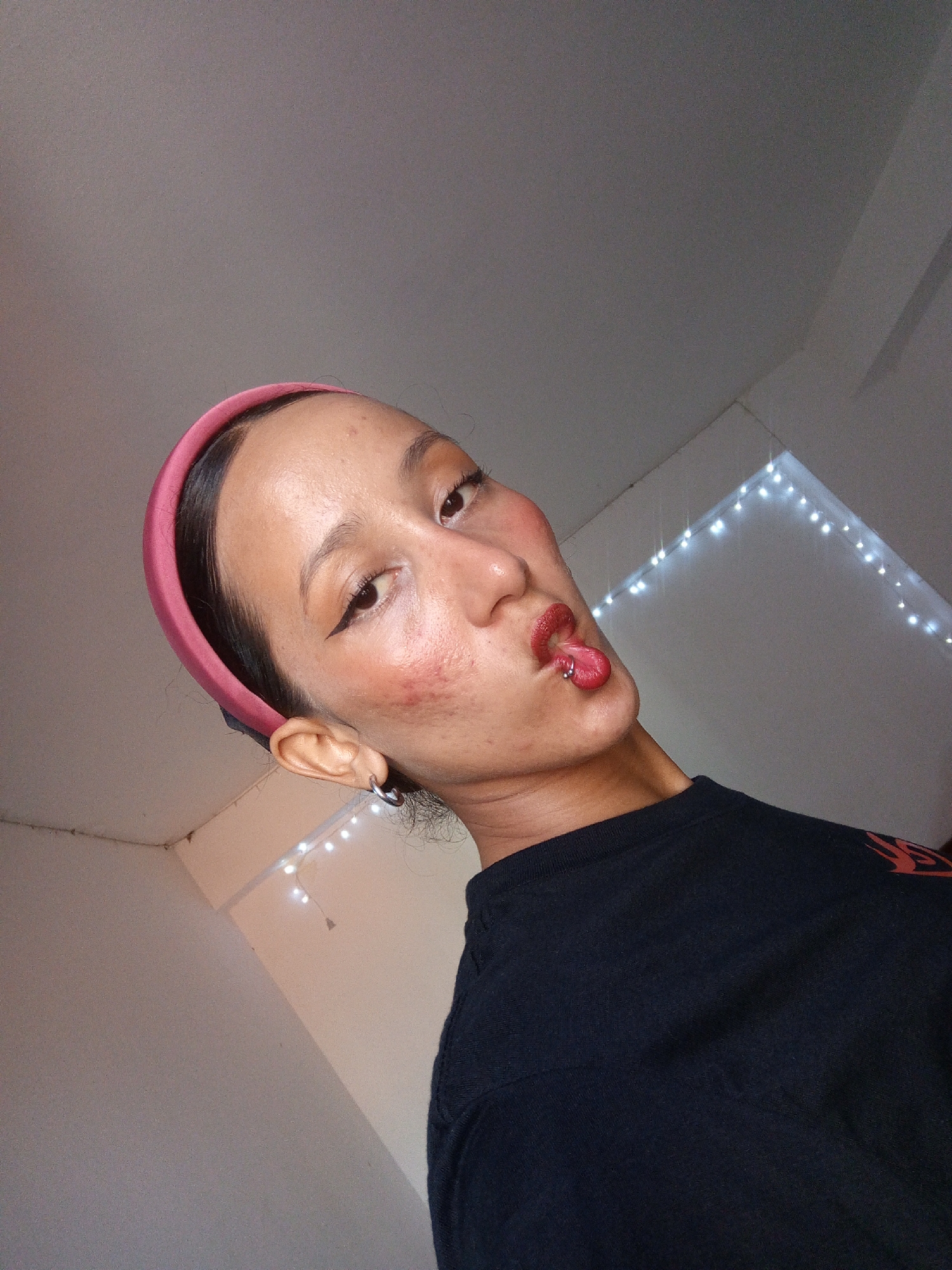 A person with dark hair pulled back by a pink headband is looking at the camera. They are wearing a black t-shirt and have a tongue piercing. The background is a white wall with string lights. The location is unknown.