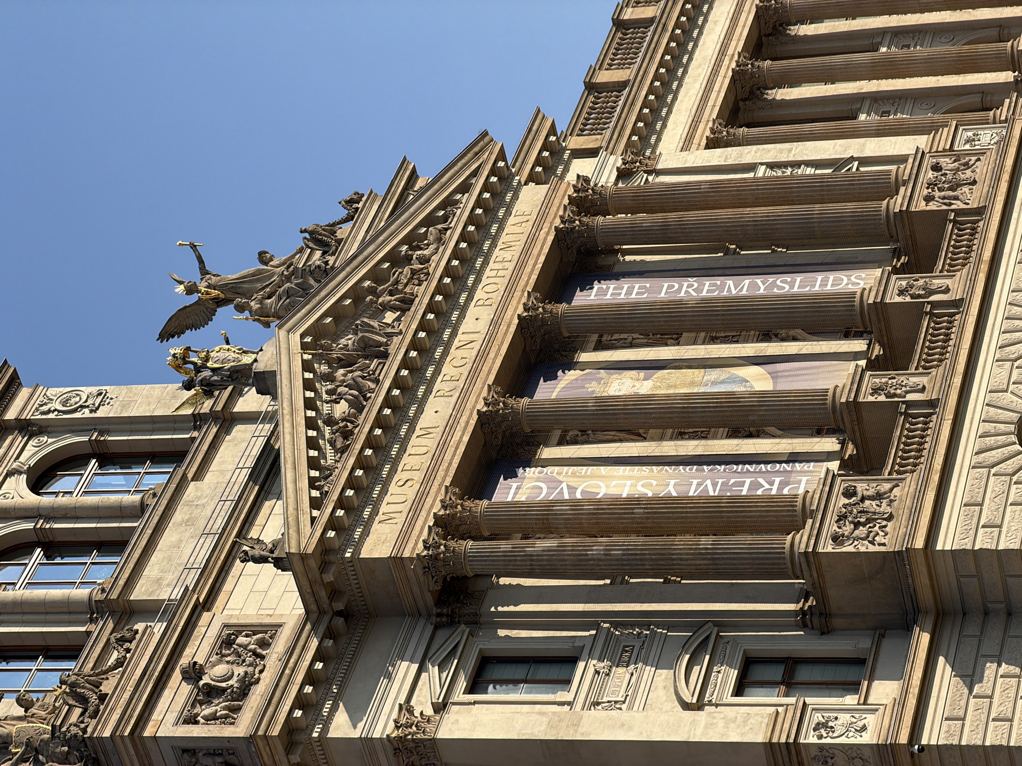 The image shows the National Museum in Prague, Czechia, on a clear day with a bright blue sky. The museum is a grand neoclassical building with ornate architectural details. In the foreground, large Corinthian columns support a portico. Banners hang between the columns, one of which displays the text "PŘEMYSLOVSCI" and "THE PREMYSLIDS". Above the portico, there is a pediment adorned with sculptures and further architectural ornamentation. Statues of winged figures and mythological scenes are visible on the roofline. Sunlight casts shadows, highlighting the three-dimensional qualities of the building's facade. The lower portion of the image shows a section of the museum's lower facade with windows and decorative carvings.