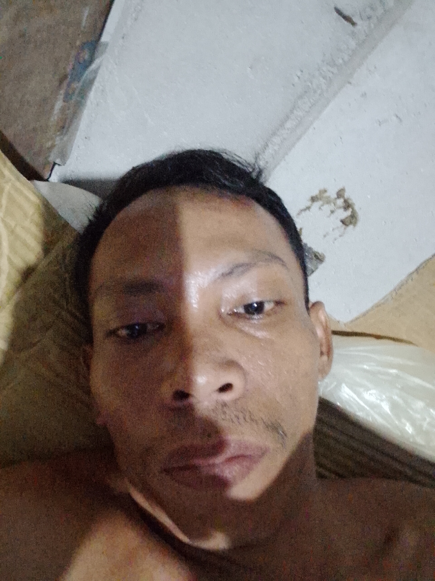 The image is a close-up selfie of an adult male with dark hair. His face is partially illuminated, with a distinct line of shadow running vertically down the center. He has dark eyes, a prominent nose, and a thin mustache. The background includes a pillow or cushion to his left and what appears to be a wall or ceiling above him, showing signs of discoloration or damage. The setting appears to be indoors. There is no specific indication of the city of Bukit Ketok, Indonesia, or evidence to determine that the photo was taken there.