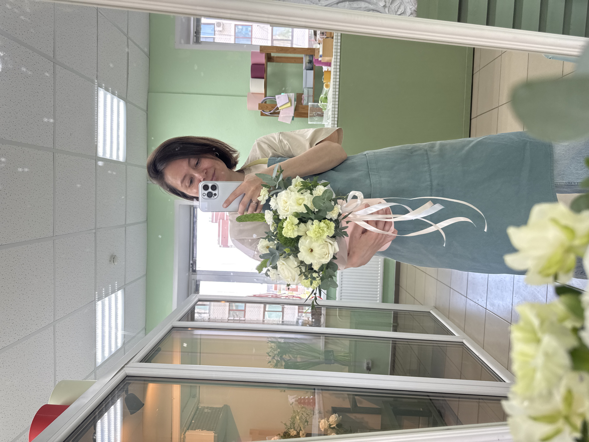 A person with dark hair and a light complexion is taking a selfie in a mirror while holding a bouquet of white flowers. The person is wearing a light-colored shirt and a blue-green apron. The bouquet is tied with light pink and white ribbons. The background shows the interior of a flower shop or a similar retail space. Shelves with floral supplies and decorations are visible. Outside the window, there is an urban scene with buildings. Based on geotagging data, the location is likely in Izhevsk, Russia. The image shows a mirror reflecting the woman holding flowers.