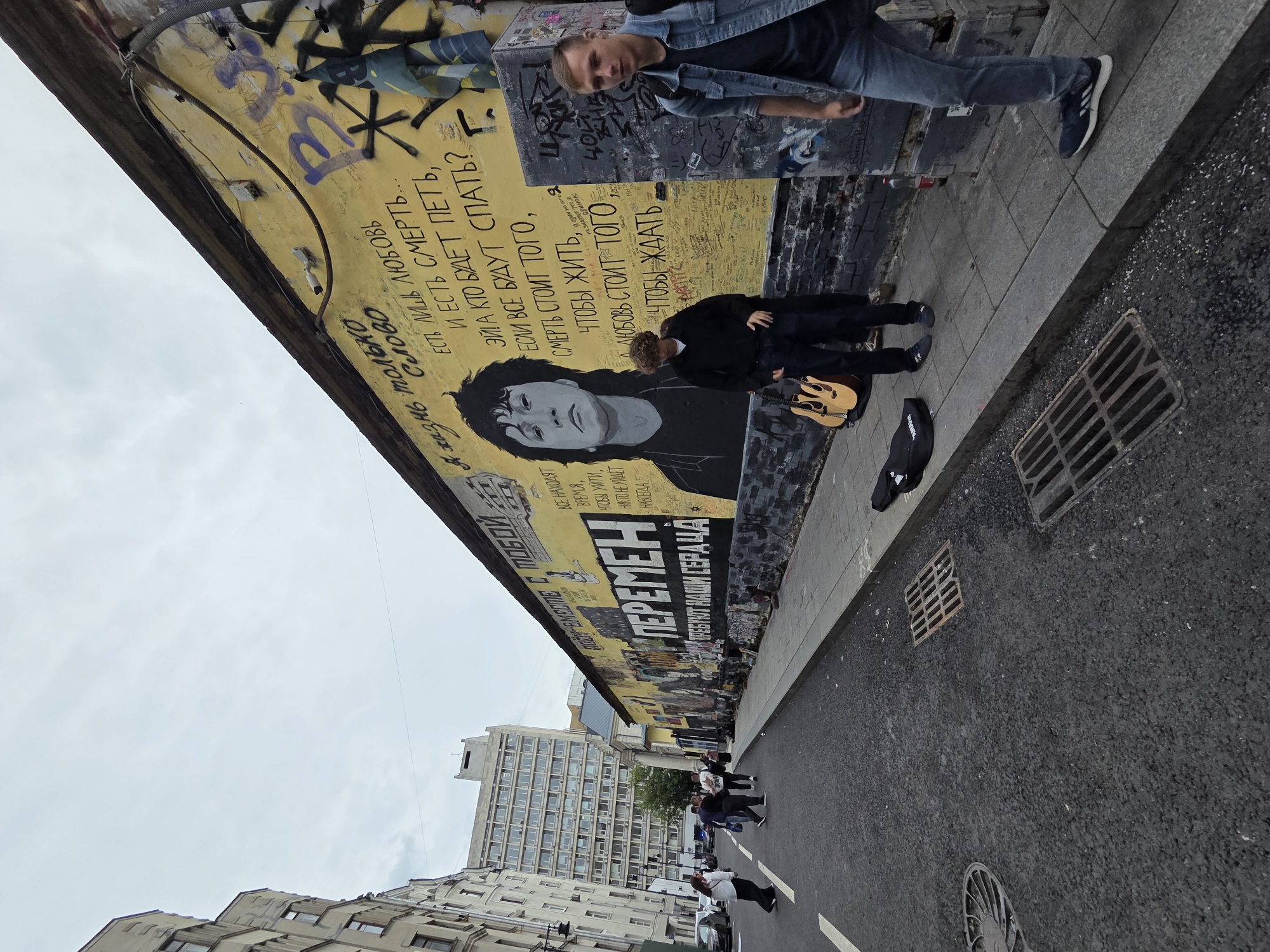 The image depicts a street scene in Moscow, Russia, specifically in the vicinity of the Tretyakov Gallery. A large mural of Viktor Tsoi, a prominent Soviet rock musician, adorns a yellow wall, accompanied by lyrics from his songs. Two young men are present near the mural. One, wearing a black hoodie and dark pants, stands beside an acoustic guitar case and a guitar. The other, in a denim jacket and blue jeans, walks past the mural, looking towards the camera. In the background, several people are walking on the sidewalk, and cars are parked along the street. The architecture suggests a historic urban setting with multi-story buildings. The overall atmosphere is that of a public space where art and everyday life intersect.