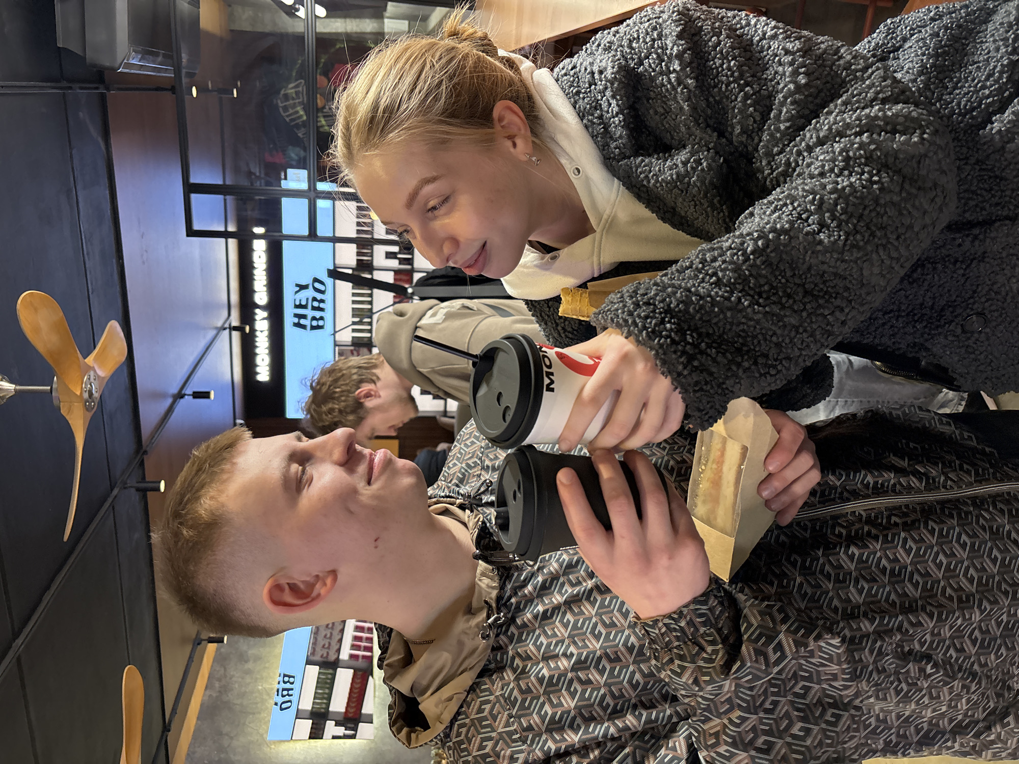 The image captures a candid moment between a young woman and a young man in what appears to be a modern cafe or restaurant. The woman, with blonde hair tied back, is on the right, leaning in and looking down at the man with a gentle expression. She is wearing a white hooded sweatshirt under a dark grey, textured fluffy jacket and holds a white coffee cup with a red and black logo (partially visible "MON").
The young man, on the left, has short, light-brown hair and is looking up at the woman. He is dressed in a dark jacket with a repeating geometric pattern in brown, beige, and black. He holds a black reusable coffee cup in one hand and a piece of food, possibly a pastry or sandwich, wrapped in a brown paper sleeve, in the other. Another person is dimly visible in the background, out of focus.
The setting is indoors, characterized by dark walls, modern ceiling lights, and what looks like a decorative wooden propeller on the wall. Visible digital signage in the background displays text, including "MONKEY GRINDE" and "HEY BRO," and a smaller sign below reads "BBQ." The overall atmosphere is casual and relaxed, suggesting a friendly interaction or shared meal. The location context is Perm, Russia.
