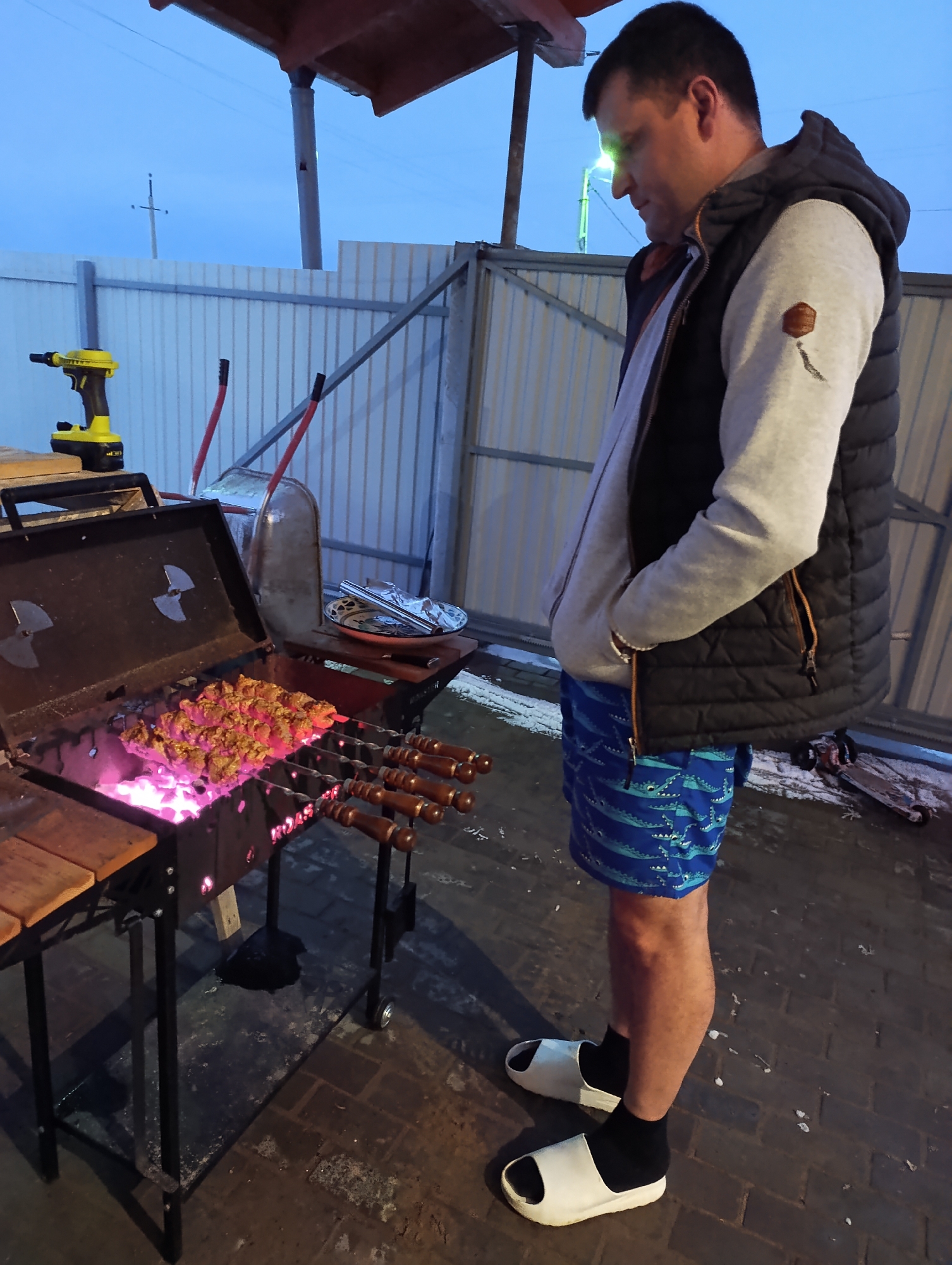 The image depicts a man standing outdoors next to a barbecue grill. He is wearing a gray hooded sweatshirt, a black quilted vest, blue swim trunks with a crocodile pattern, black socks, and white slide sandals. The grill is open, and meat is cooking on skewers over glowing coals. To the left of the grill, a yellow power drill sits on a wooden surface. Behind the grill, there is a metal wheelbarrow and a plate covered with foil. The background shows a corrugated metal fence and a light pole, suggesting an urban or suburban setting. The location is Chaikovsky, Russia, based on visual cues like the architecture and overall atmosphere. The man appears to be checking on the food cooking on the grill. The scene is somewhat informal, suggested by the clothing and the setting.