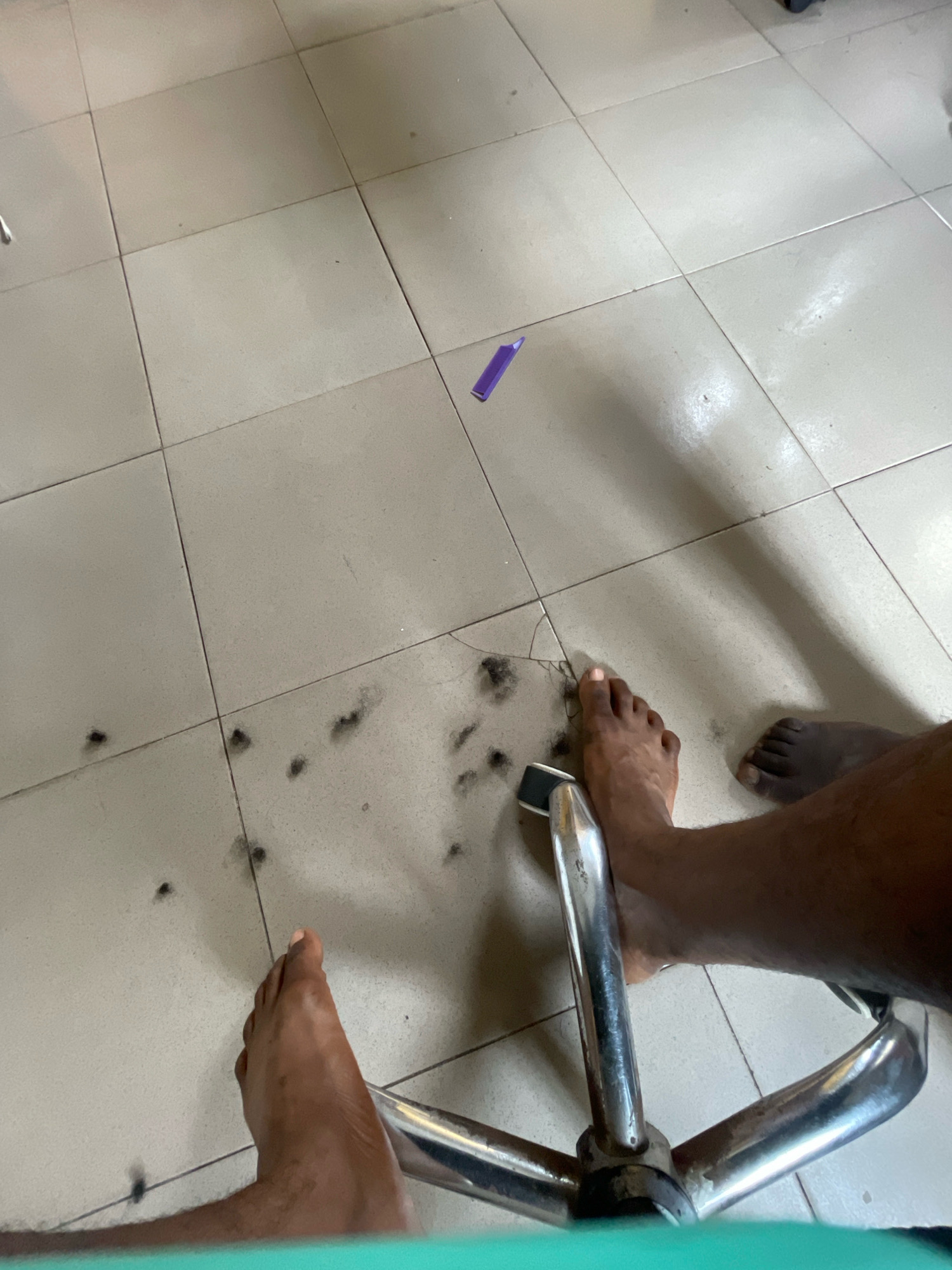 The image captures a close-up view of a tiled floor in Ado, Nigeria, with scattered hair clippings and a single purple object, possibly a crayon or pen cap. The foreground is dominated by the lower legs and feet of at least two individuals, one of whom is positioned next to a chrome-legged chair. The lighting suggests an indoor setting, possibly a home or a barber shop, given the presence of hair trimmings. The overall scene conveys a sense of recent activity, likely a haircut or grooming session, interrupted or completed.