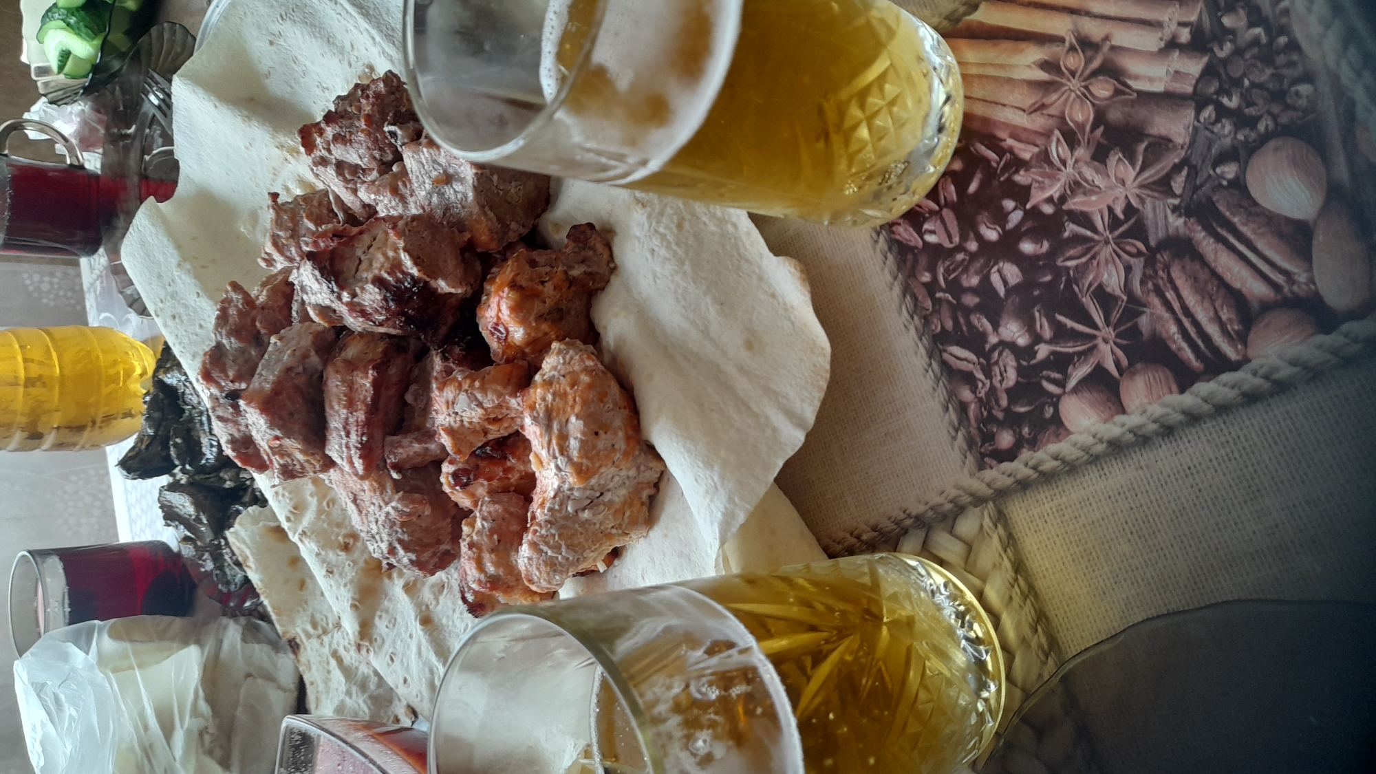 The image depicts a close-up, overhead view of a table laden with food and drinks, likely in a domestic setting in Bagayevskaya, Russia. The focal point is a platter of cooked meat, appearing to be shashlik or kebabs, served on a bed of flatbread. Two glasses filled with a pale yellow, effervescent beverage, presumably beer, are prominent in the foreground. To the left, a glass of dark red liquid, possibly wine or juice, is visible alongside a partially opened plastic bag. In the background, there are other containers with food items, including what looks like a bowl of sliced cucumbers. The table surface is covered with a patterned cloth featuring images of coffee beans and spices. The lighting suggests an indoor environment, possibly during daylight hours. There are no individuals visible in the image.