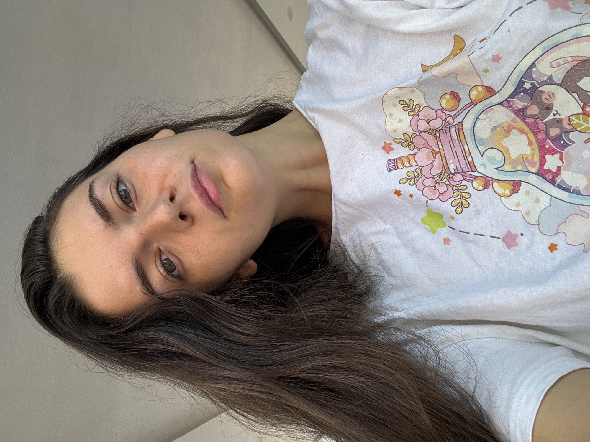 A woman is seen from a tilted, close-up perspective, wearing a white t-shirt with a cartoon illustration of a decorated glass bottle. She has long, brown hair, fair skin, and green eyes. She is indoors against a neutral colored wall. The location in Mũi Né, Vietnam, cannot be determined from the provided image.