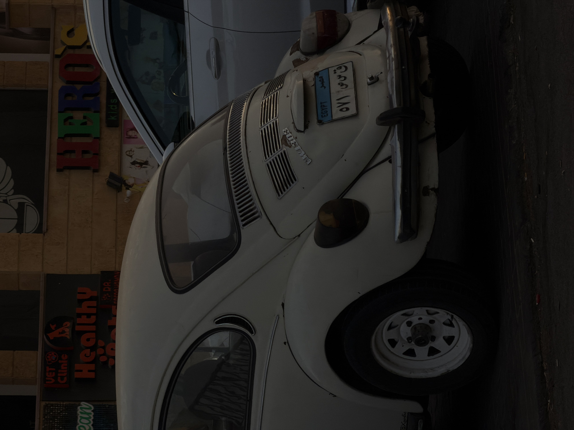 A vintage white Volkswagen Beetle is parked on a street in Sharm El Sheikh, Egypt. The car is seen from the rear, and its license plate is visible. Behind the Beetle, a modern silver SUV is also parked. In the background, there are storefronts with signs. One sign reads "HERO'S KIDS," and another has "VET CLINIC" and "Healthy Pets." The lighting suggests it is either early morning or late afternoon, casting long shadows. The overall scene depicts a typical urban street with a mix of old and new vehicles and commercial establishments.