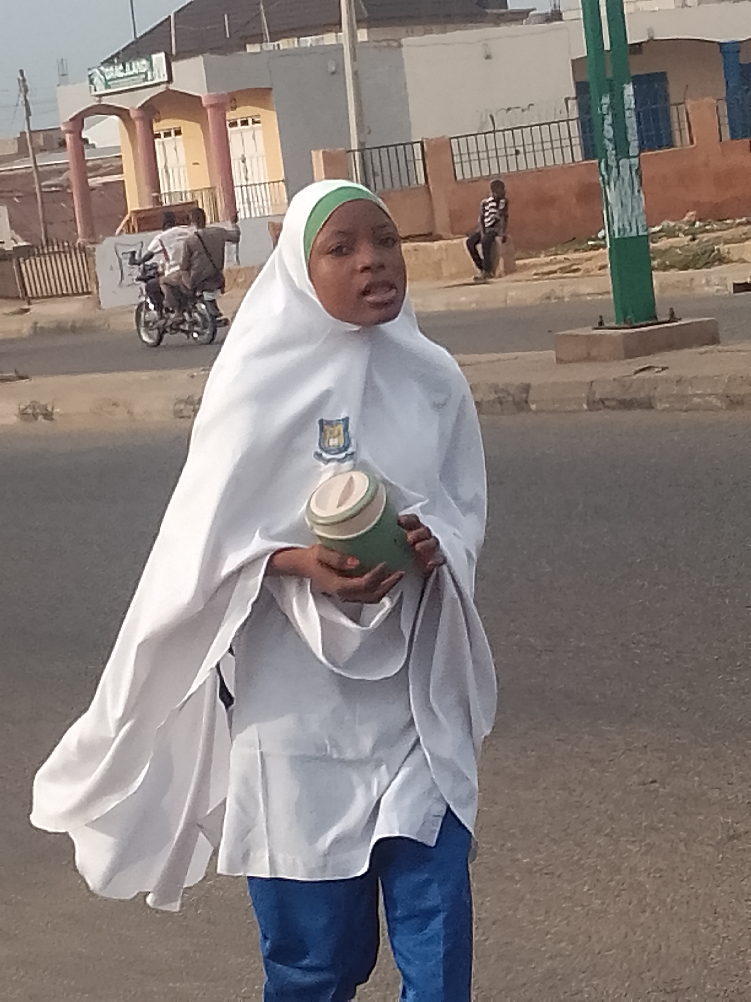 In Arawa, Nigeria, a young person wearing a white hijab and a white uniform walks on a street. The person is holding a cylindrical object with green and tan accents. In the background, a motorcycle with two occupants is visible, and further in the distance, a building with pillars and arched entrances stands. A green pole with white markings is also present in the scene.