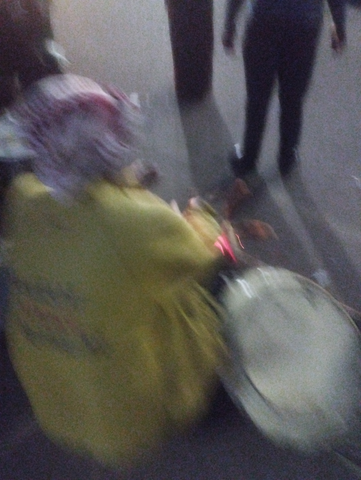 The image shows a blurry, low-light scene that appears to be outdoors, possibly at night or in dim conditions. In the foreground, a person is visible wearing a bright yellow garment, with what looks like purple or reddish hair, suggesting they might be female. Their body is blurred, indicating movement or a long exposure. To the right, a large, reflective, circular object is present, possibly a vehicle wheel or a reflective surface. In the background, another person is seen standing, wearing dark clothing. Their legs and lower torso are discernible. The environment is indistinct but appears to be a paved surface, hinting at a street or walkway. The overall impression is of a candid, possibly candid shot captured in a dynamic urban setting. Without further context or clearer details, it is not possible to definitively identify the location as Jos, Nigeria.