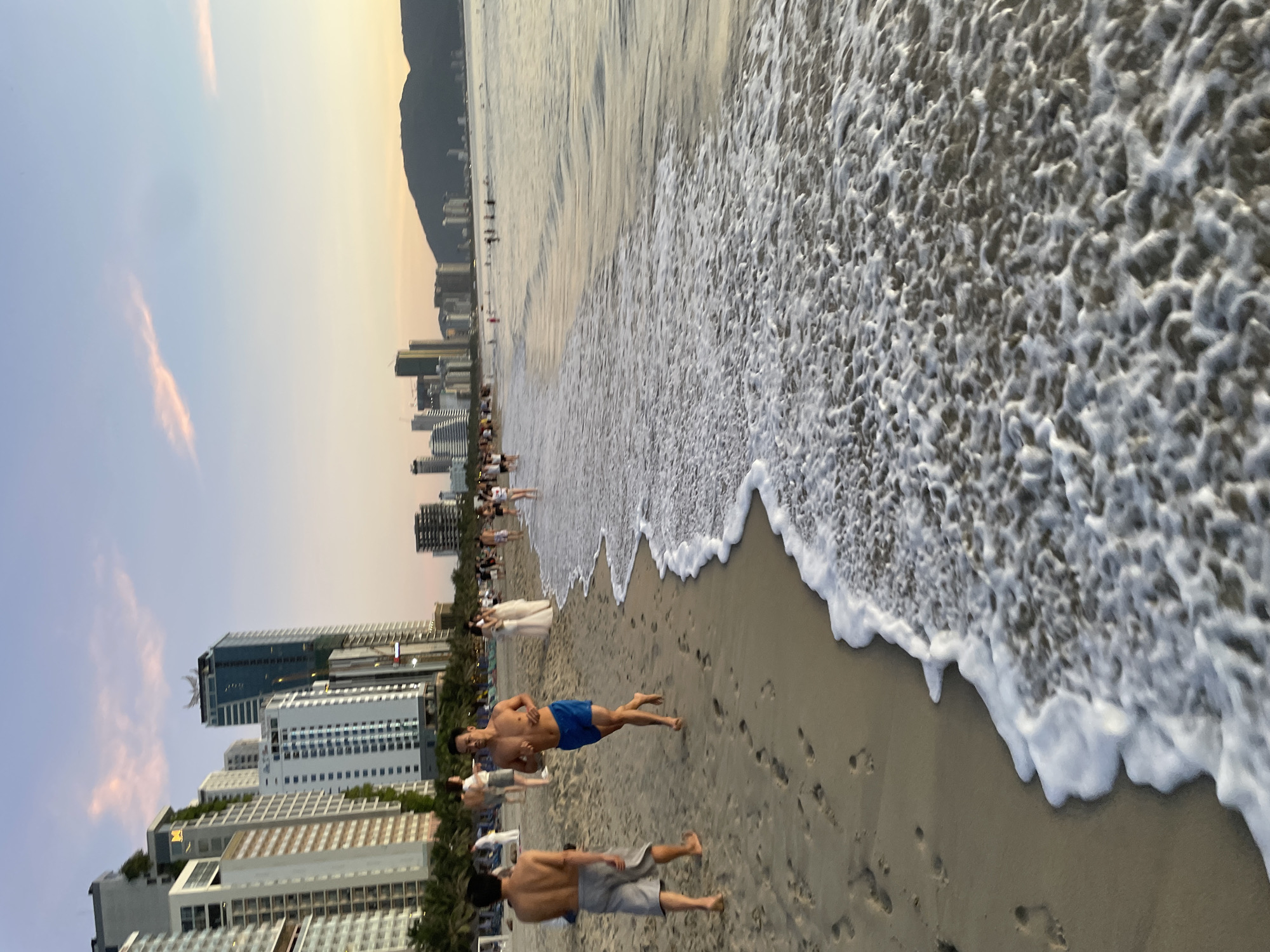 The image shows a beach scene in Da Nang, Vietnam, with the cityscape in the background. The sky is a pale blue with a few wispy pink clouds, suggesting it is either sunrise or sunset. The water is calm and shallow, with gentle waves lapping at the shore, creating a foamy white edge on the wet sand. In the foreground, two men are visible on the beach. One man on the left is walking away from the camera, wearing gray shorts. The man on the right is running towards the left, wearing blue swim trunks and appears to be shirtless. Further down the beach, a crowd of people are visible, some walking, some standing, and some in the shallow water. The background is dominated by a line of modern, tall buildings, characteristic of a coastal city like Da Nang. Palm trees are also sparsely scattered along the beachfront. The overall impression is of a relaxed and active beach environment during the golden hour.