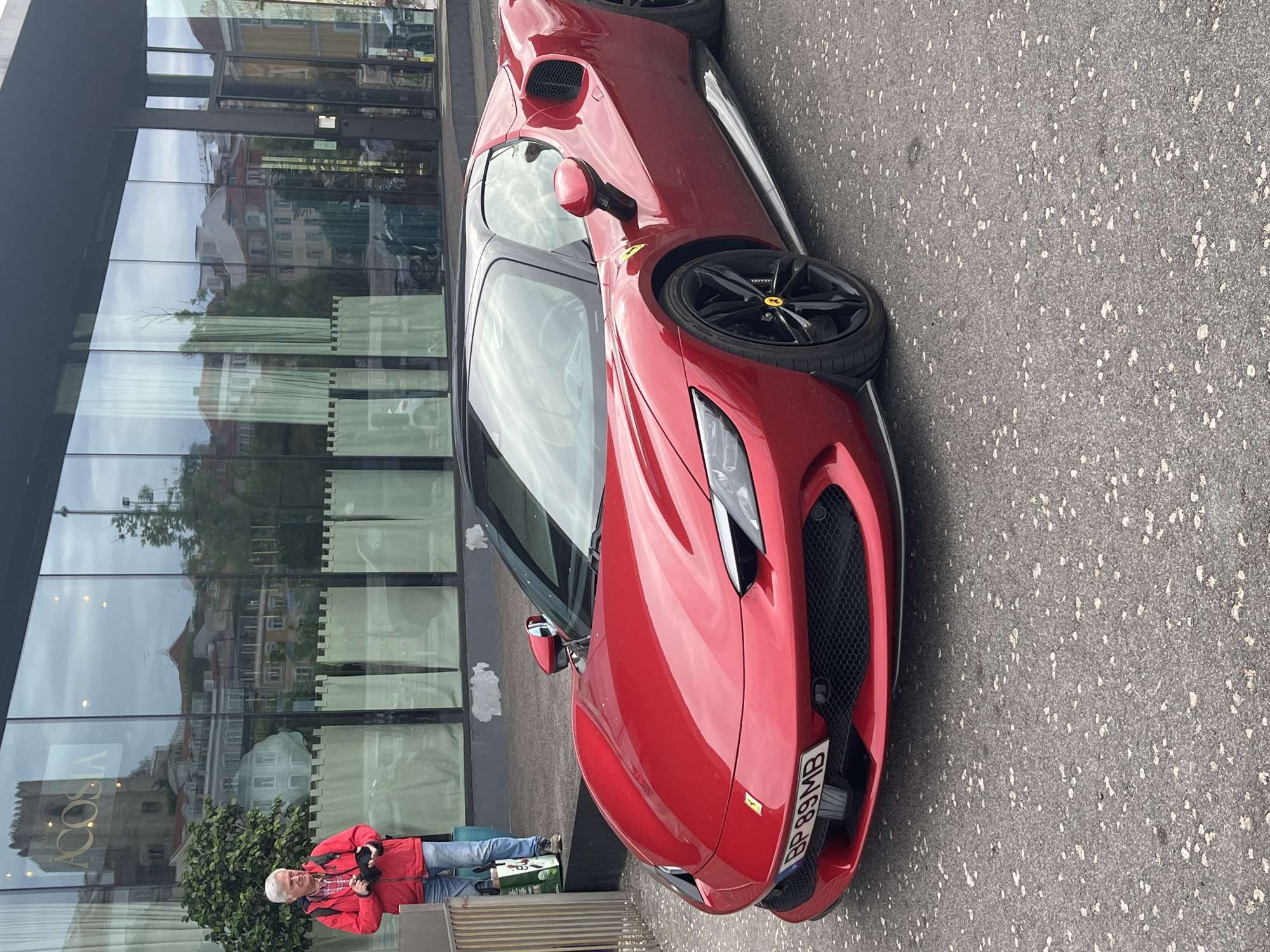 A red Ferrari sports car is parked on a paved street. The car's front end is facing right. A man in a red jacket stands on the left side of the frame, appearing to take a photo of the car. The car is parked in front of a modern building with reflective glass windows that mirror the surrounding cityscape. The car's license plate is visible. The license plate appears to be a European plate, but the country is impossible to determine. The license plate reads "BP 89MB". The location appears to be Lisbon, Portugal, based on the reflected cityscape in the building's windows. The paved street is gray and textured. The man has gray hair and is wearing jeans.