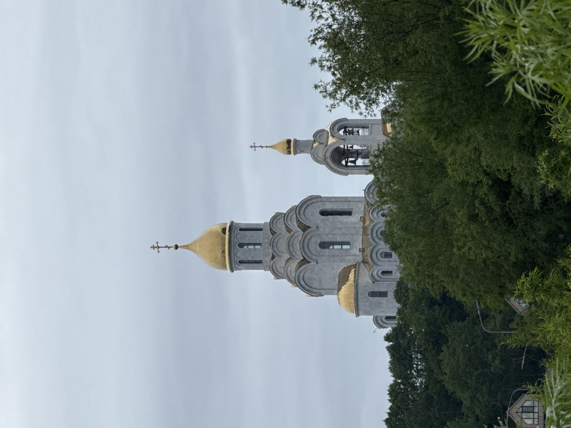 The image depicts a large, light-colored Orthodox church with golden domes, positioned on a hill and partially obscured by lush green trees. The sky is overcast and gray. To the left of the main church structure, a smaller bell tower with bells is visible. In the foreground, dense foliage and trees dominate the lower half of the frame. Two small structures with triangular roofs and windows are nestled amongst the trees in the lower left and right portions of the image, along with streetlights. The overall scene suggests a somewhat remote or park-like setting within or on the outskirts of a city. This church is the Church of the Resurrection of Christ, located in Petropavlovsk-Kamchatsky, Russia.