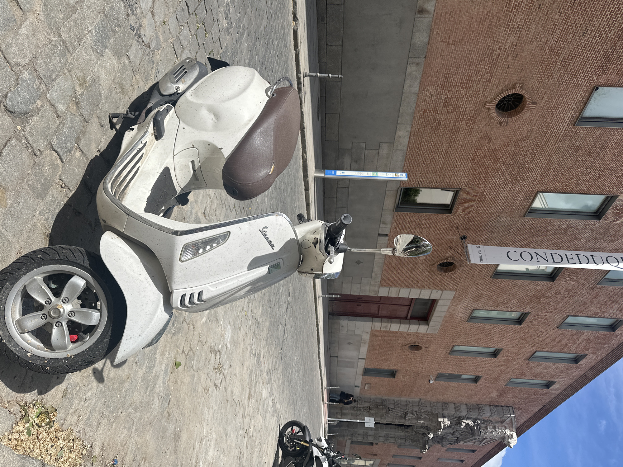The image shows a white Vespa scooter parked on a cobblestone street in Madrid, Spain, based on the "CONDE DUQUE MADRID" sign visible on a building in the background. The building is made of red brick with several rectangular windows. The sky is blue. Another parked motorcycle is visible in the distance. The Vespa has a brown seat and appears to be dusty.