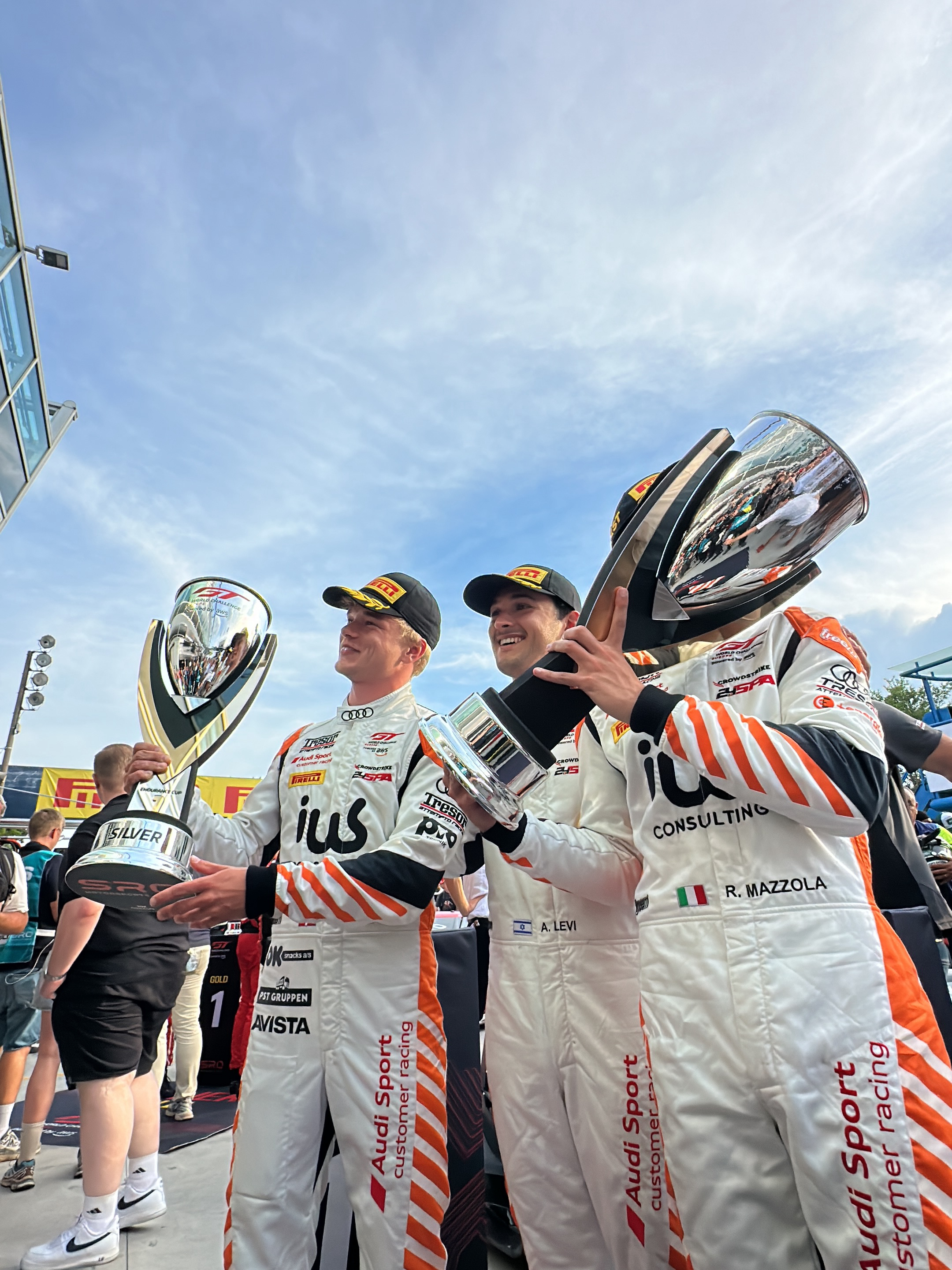 The image shows three men in racing suits standing together and holding trophies. They appear to be celebrating a victory, as they are smiling and holding their trophies up. They are wearing matching white racing suits with orange stripes and black caps. The suits have logos for Audi Sport customer racing, ius Consulting, and Tresor. The man on the left is holding a silver trophy and the man in the middle has the Israeli flag on his suit, and the man on the right has the Italian flag on his suit. The background shows a blue sky with white clouds. The location is likely in Monza, Italy, as the man on the right has the Italian flag on his suit.