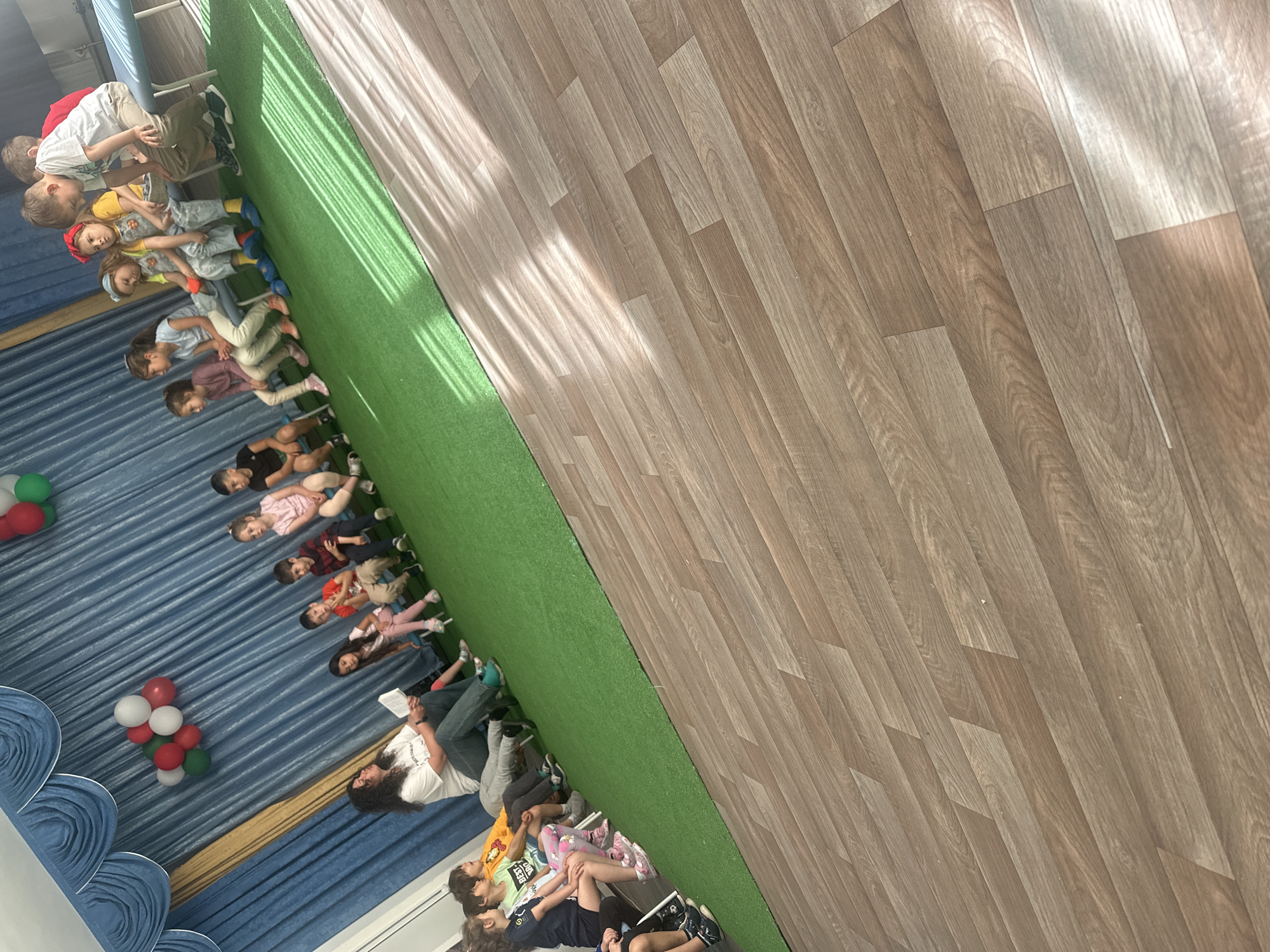 A medium-shot, eye-level indoor view shows a group of children sitting on a blue rug, likely in a classroom or educational space. A woman is seated among the children, seemingly reading to them from a book. The room's floor transitions from the blue rug to a strip of green artificial turf and then to wood laminate flooring. A cluster of red, white, and green balloons stands near the left edge of the frame. Given the colors and classroom setting, this photo may be from a school in Plovdiv, Bulgaria.