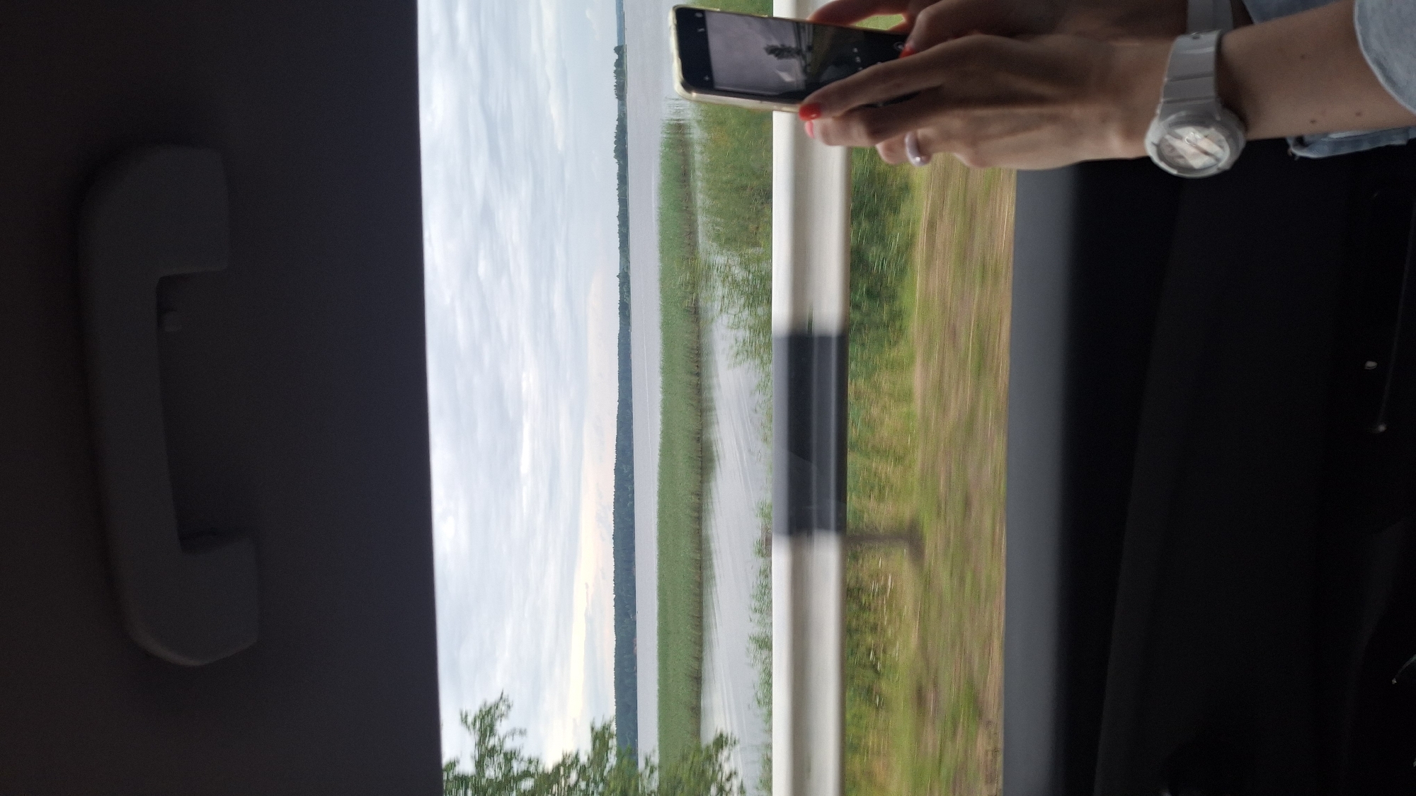 In this image, taken from inside a vehicle, a person's hands are holding a smartphone to capture a photograph of a serene natural landscape. The scene is a view from a car window looking out at a body of water, likely a lake, bordered by a dense line of trees in the distance. In the foreground, the hands, adorned with a white wristwatch and a ring, are positioned to frame the shot on the phone screen. The phone itself displays a reflection of the surroundings, including a tree and the sky. The environment outside the car suggests a rural or semi-rural setting, possibly near Ilyanskiy selskiy Sovet, Belarus, given the presence of water and trees. The overall mood is one of observation and capturing a moment of travel.