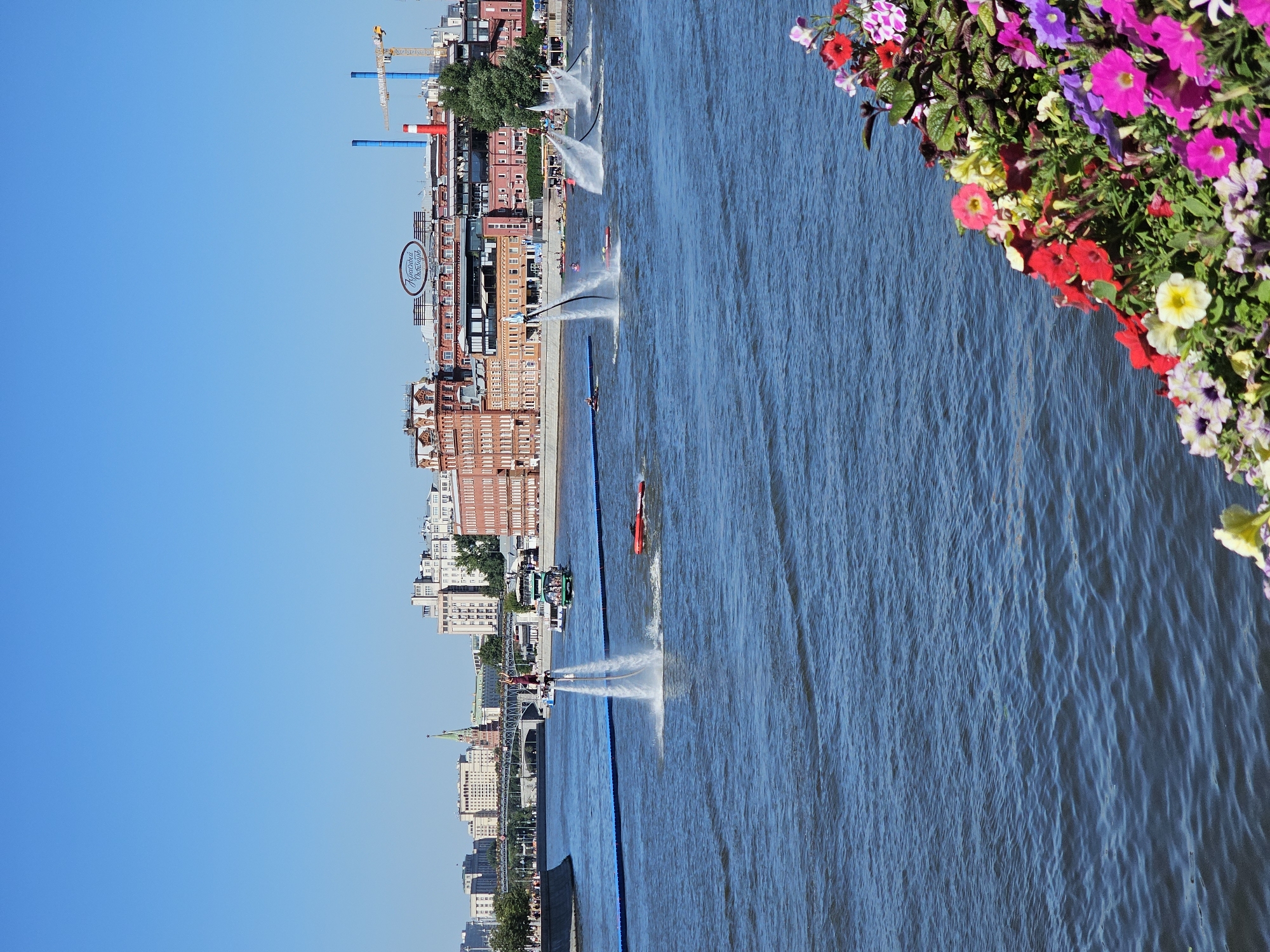 In Moscow, Russia, on a clear day, the Moskva River flows through the city. In the foreground, a vibrant cluster of colorful flowers, including red, pink, purple, and yellow petunias and other blooms, lines the edge of the water. The river is a dynamic scene of activity, with multiple individuals engaged in flyboarding. One person is prominently positioned in the center, suspended mid-air by powerful jets of water from a device attached to their feet, with streams of water erupting around them. To the right, two other flyboarders are also airborne, creating plumes of water spray. Further in the distance, a red jet ski glides across the water. A tour boat, filled with passengers, travels along the river, and a smaller vessel is also visible. The riverbanks are lined with a curved wall, and beyond that, the cityscape of Moscow unfolds. Prominent buildings, including a large red brick structure with a distinct oval sign bearing Russian text, are visible. The city skyline is punctuated by other urban structures, and a construction crane is visible in the background. The sky is a clear, bright blue, indicating fair weather.