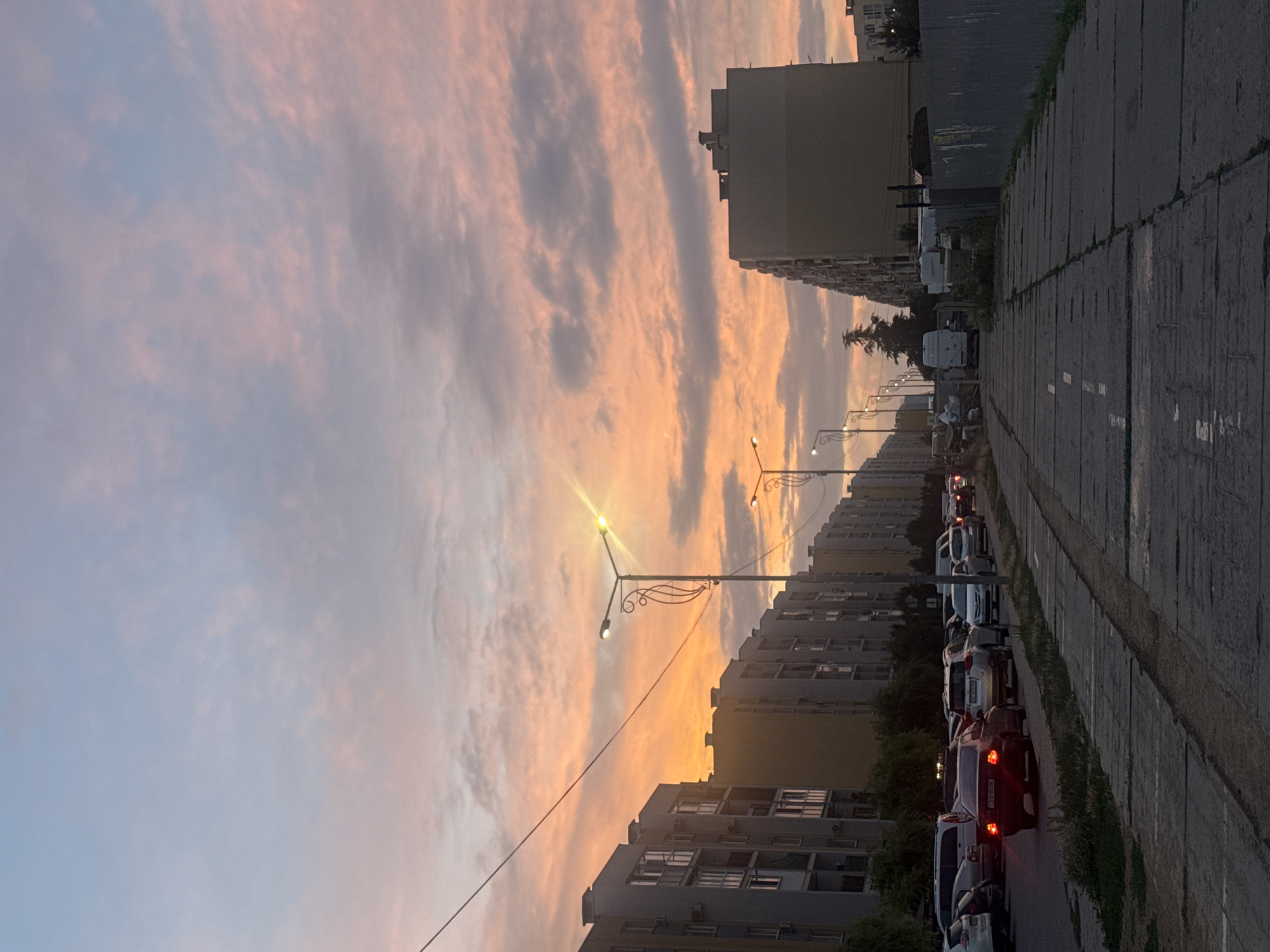 The image depicts a street scene at dusk in Avanhard, Ukraine. Tall, apartment buildings line both sides of the street, with their facades illuminated by the warm hues of the setting sun. A line of cars is parked and in motion along the street. The sky is filled with dramatic clouds displaying shades of orange, pink, and blue. Streetlights are visible, with one in the foreground emitting a bright yellow glow. A paved pedestrian walkway runs alongside the street. In the background, a fence and what appears to be construction materials are present. The overall impression is that of an urban environment transitioning from day to night under a colorful sky.