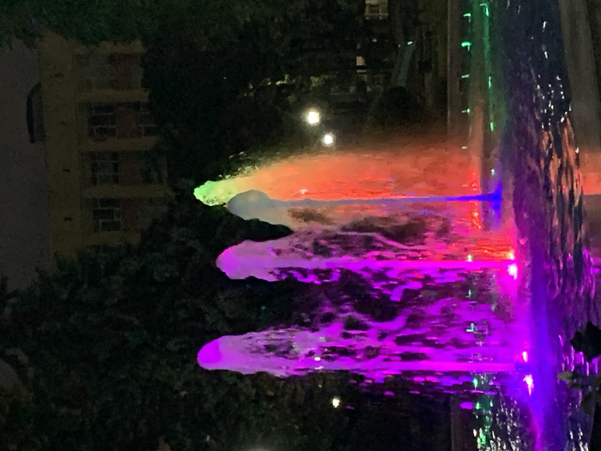 The image depicts a night scene of a brightly lit fountain in a city park. The fountain features several streams of water illuminated by vibrant purple and red lights, with one stream transitioning to green at the top. The water appears to be in motion, creating a dynamic effect. In the background, there are dark trees and a building with a yellowish facade, illuminated by windows. Several streetlights are visible, emitting a soft glow. The ground around the fountain is wet, reflecting the colorful lights. The overall impression is that of a serene and visually appealing urban landscape at night.