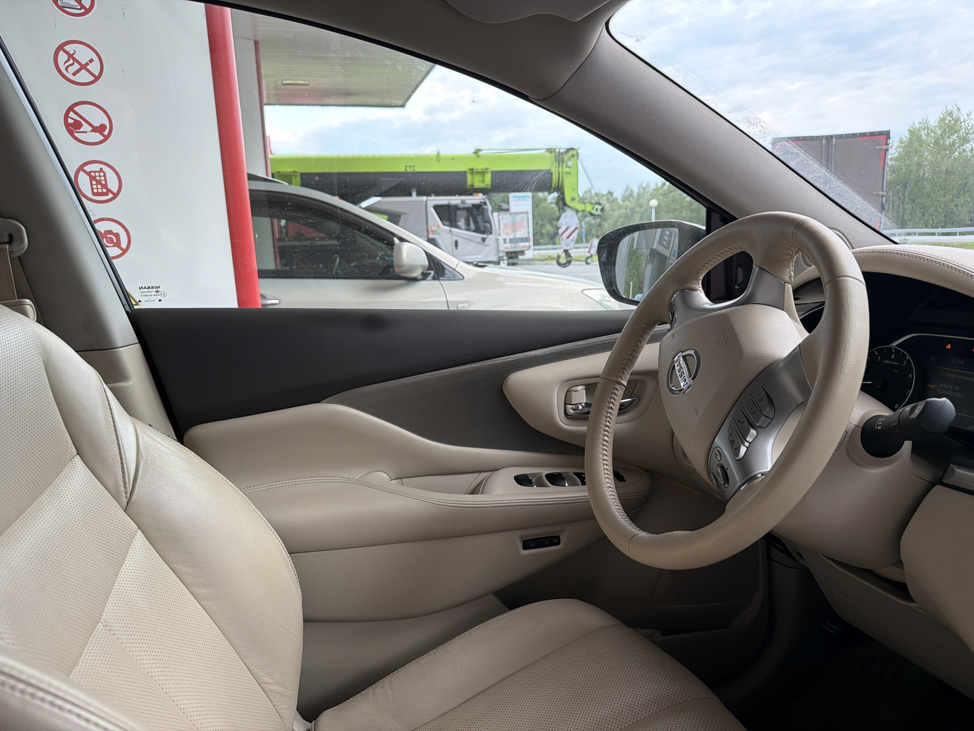 The image shows the interior of a car, likely a Nissan, viewed from the driver's seat. The car is parked at a gas station, evidenced by the fuel pump canopy visible through the windshield. Outside the car, a large green construction crane and a truck are visible, along with trees and a cloudy sky. The car's interior features beige leather seats, a leather-wrapped steering wheel with controls, and a dashboard with a digital display. There are also several red circular warning signs on the white pillar of the gas station canopy, indicating prohibitions against smoking, using mobile phones, and other activities. The scene appears to be a typical day at a roadside service area, with the car likely stopped for refueling or a break. The location is identified as Shchetkovo, Russia.