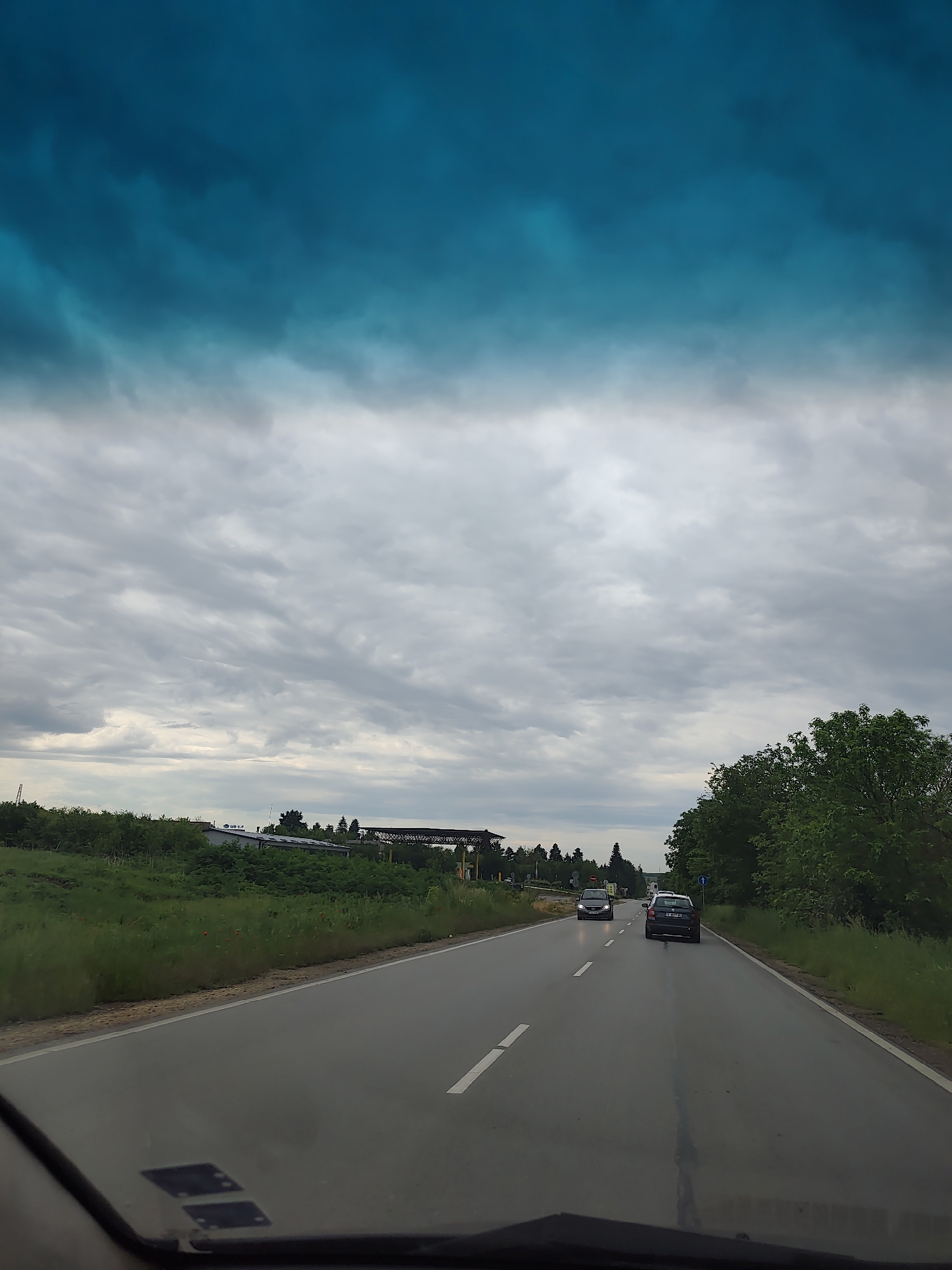 The image depicts a road in Dobrich, Bulgaria, viewed from inside a car. The sky is predominantly overcast with grey and white clouds, partially obscured by a blue-tinted windshield. The road is wet, with a solid white line on the right side and a broken white line dividing the lanes. Two cars are visible on the road ahead: one is grey, further ahead in the opposite lane, and the other is a black car that is a shorter distance in front, in the same lane. Green foliage lines both sides of the road. A building complex is visible in the distance to the left.