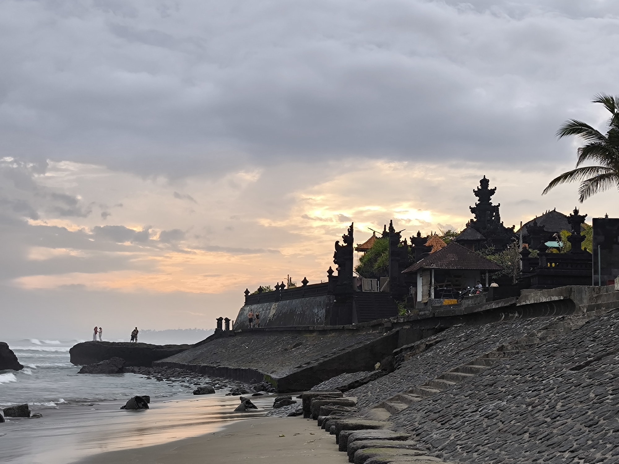 This is a photograph of Tanah Lot Temple, a sea temple on a rock formation in North Kuta, Bali, Indonesia, at sunset. The sky is filled with dramatic, overcast clouds that are tinged with the orange and pink hues of the setting sun. The ocean is choppy, with waves crashing against the rocky shore. In the foreground, a sandy beach is visible, with scattered rocks. Along the coastline, a stone wall and steps lead up to the temple complex, which is characterized by its dark, intricately carved structures and tiered rooftops. A few people are visible on the rocks near the water's edge, and others are near the temple entrance. A palm tree frond hangs into the frame from the upper right. The overall atmosphere is one of dramatic natural beauty and cultural significance.