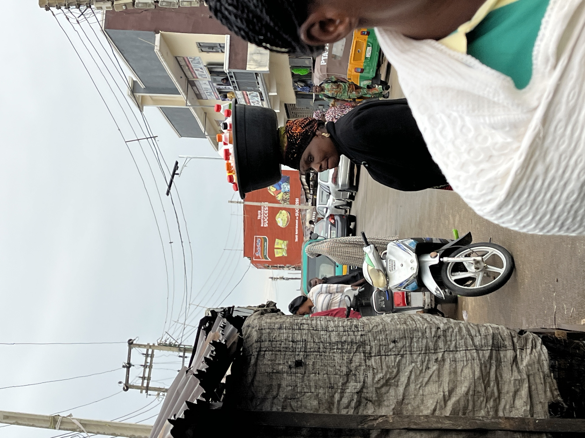 The image captures a street scene in Jos, Nigeria. An African woman wearing a patterned headscarf and black top balances a black tray filled with small, brightly colored items, possibly drinks, on her head. She is walking on the side of a street, with a white shawl draped over her shoulder, near the top right of the image.
In the background, there are vehicles, including a motorcycle, a car, and a yellow and green auto rickshaw (Keke Napep). A large advertisement for Indomie noodles is visible.
The foreground features a covered structure, potentially a stall or temporary shelter, constructed with corrugated metal and a gray fabric covering. Utility poles and electrical wires are visible overhead. Several other people are on the street.