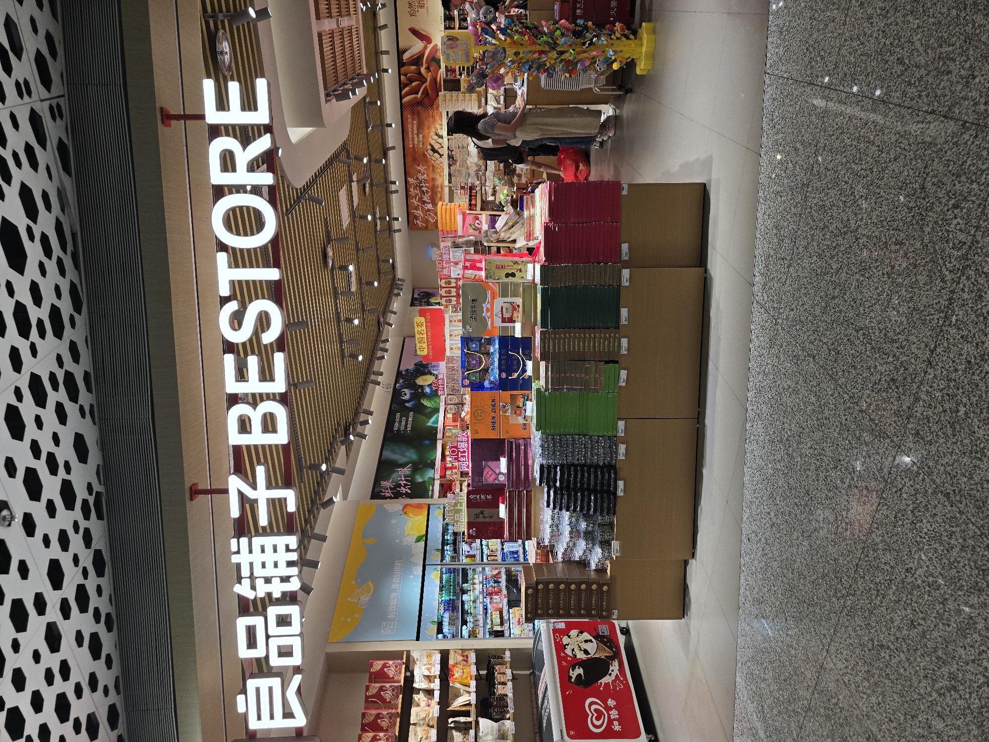 The image shows the interior of a Bestore shop in Bao'an District, China. The store is well-lit and organized, with shelves stocked with various snacks and confectionery products. A prominent sign above the entrance reads "良品铺子 BESTORE". In the foreground, there are several display stands showcasing boxes of snacks. To the left, a red freezer with an ice cream advertisement is visible. In the middle ground, customers are browsing the aisles, with one woman in a grey top and beige pants standing at a counter. The background reveals more shelves filled with products, refrigeration units, and promotional posters on the walls. The floor is a polished speckled grey tile. The ceiling features a modern design with hexagonal patterns and recessed lighting.