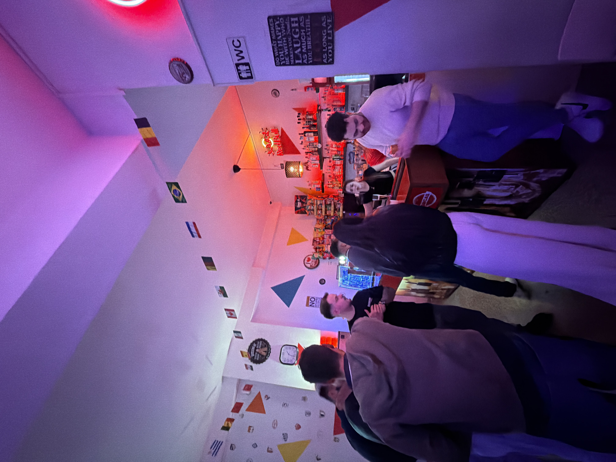 People are in a bar. The environment is a small bar with international flags and other wall decor. There's a bar with a bartender behind it and several patrons standing around. A sign reading "WC" indicates the bathroom. Another sign reads "DO WHAT MAKES YOU HAPPY, BE WITH WHO MAKES YOU SMILE. LAUGH AS MUCH AS YOU BREATHE AS LONG AS YOU LIVE." The city is Vila Real, Portugal.