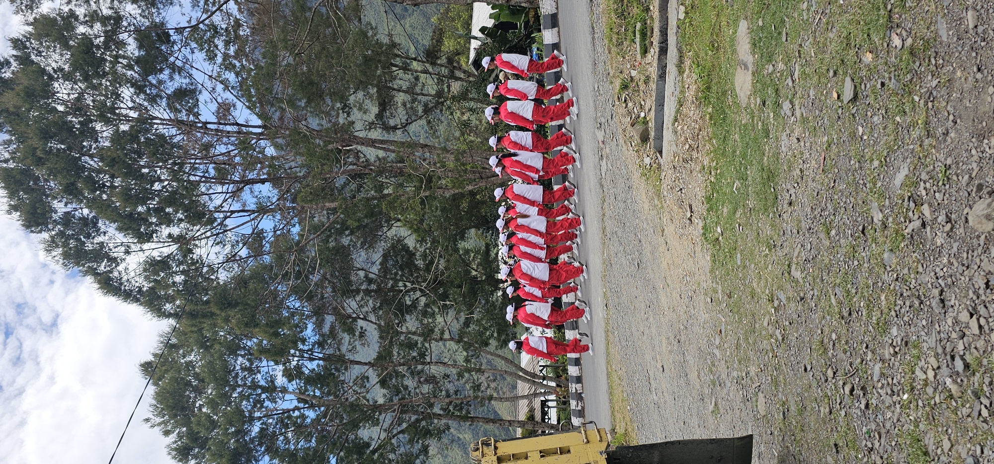 In Puncak Jaya, Indonesia, a group of individuals, likely students or participants in an organized event, are walking in a single file line along a paved road. They are all wearing matching red and white tracksuits with white caps. The group is moving from left to right in the frame. The environment is outdoors, with tall, sparse evergreen trees lining the background. The road appears to be on a slight incline, with a gravelly shoulder on the left side of the frame. In the foreground, to the far left, a portion of a large yellow piece of machinery is visible. The sky is partly cloudy with a mix of white clouds and blue sky. The terrain beyond the road is hilly and covered in dense green vegetation.