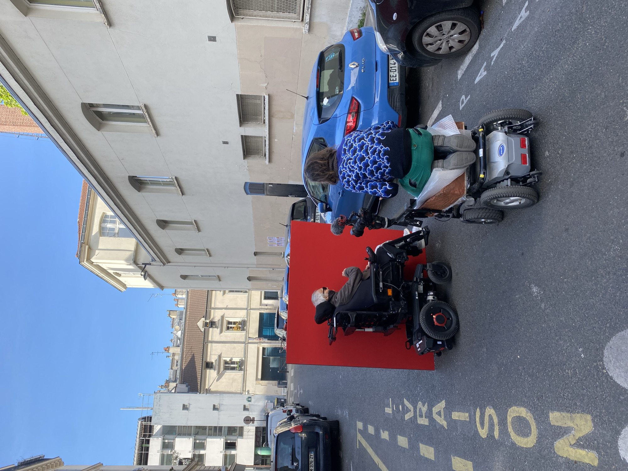 The image depicts an outdoor urban scene on a sunny day with a clear blue sky, likely in Paris, France. The perspective is looking down a street lined with buildings.
In the foreground, two individuals are positioned on a paved street featuring yellow road markings. One person, with gray hair, is seated in a black motorized wheelchair with red accents, facing left. Attached to the front of this wheelchair is a microphone on a stand, suggesting recording is taking place. Behind this individual, another person with curly brown hair, wearing a blue and white patterned top and green trousers, is bending over a second motorized wheelchair. This second wheelchair, partially obscured, has a prominent silver-grey battery pack labeled "Invacare." This person appears to be adjusting something or interacting with items placed on the wheelchair's platform, which is wrapped in a light-colored material.
A large, vivid red rectangular panel or wall is set up vertically on the street, serving as a backdrop directly behind the wheelchairs and the two individuals.
Several cars are parked along the street. Immediately behind the second person and the red panel is a blue Renault Clio with a French license plate starting "EE-014-". Another dark car is parked partially visible next to it. Further down the street to the left, more cars are parked, including a dark Peugeot, and the perspective extends towards distant buildings under the bright sky. The buildings lining the street exhibit typical urban architecture, with multiple windows and varying facades.
Visible yellow text painted on the asphalt includes "RAISON" and a partial "LIVR..." (likely "LIVRAISON," meaning delivery), along with a "P" indicating parking. The scene suggests an event, possibly a film shoot or a public demonstration, given the microphone and the prominent red panel set up on the street.