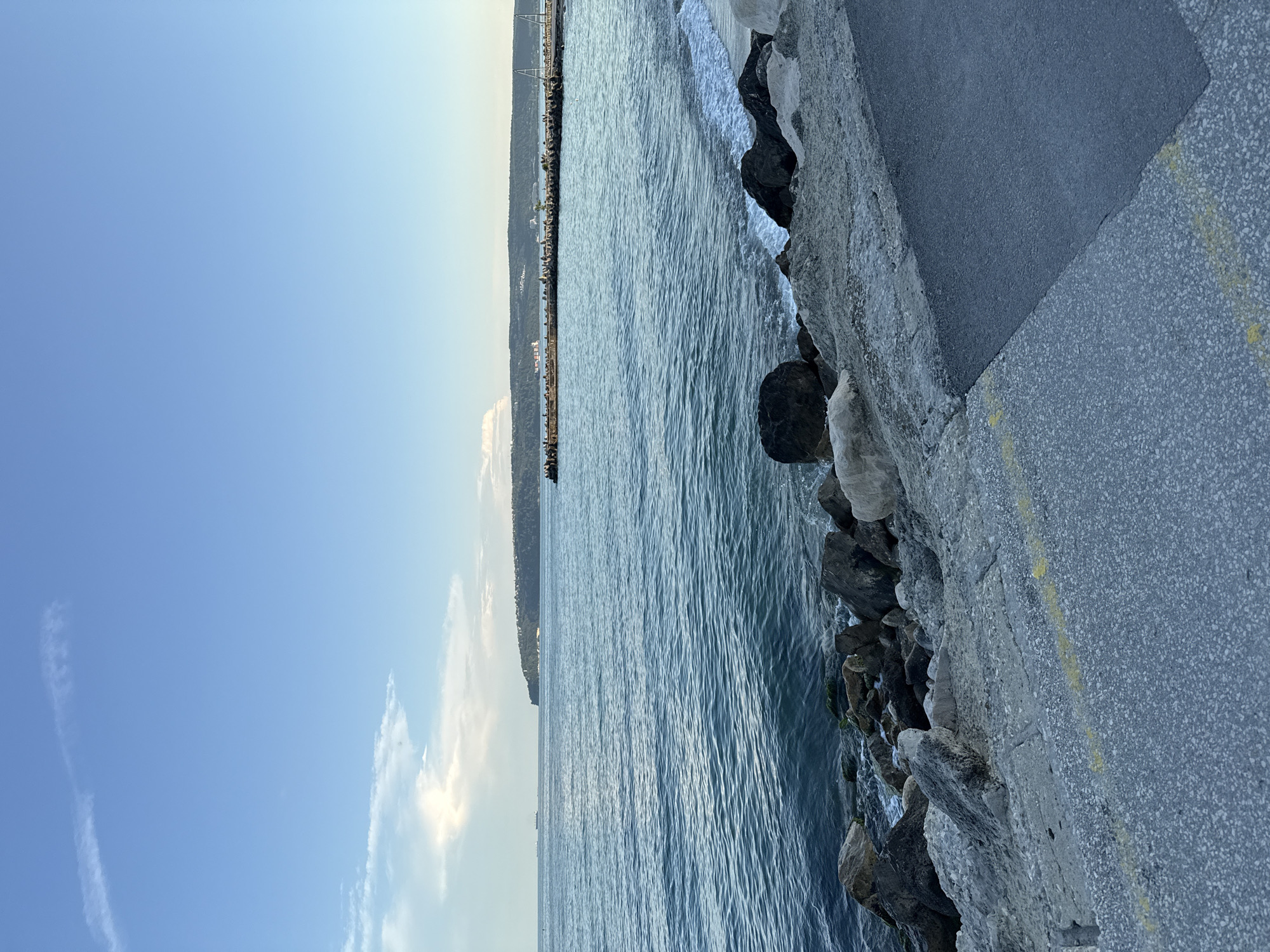 The image depicts a seascape with a rocky shoreline and a paved area in the foreground. The sea is a calm, deep blue with gentle waves lapping at the shore. Large, dark rocks are scattered along the coastline, some partially submerged in the water. In the distance, a long pier extends into the sea, lined with what appear to be people and structures. Beyond the pier, a lush, green hillside rises, capped by a few buildings. The sky is a clear, pale blue with wispy white clouds scattered near the horizon. The paved area in the foreground is a dark gray asphalt, with yellow painted lines indicating a pathway or markings. The lighting suggests it is either early morning or late afternoon, with soft, diffused light. There are no visible people in the immediate foreground.