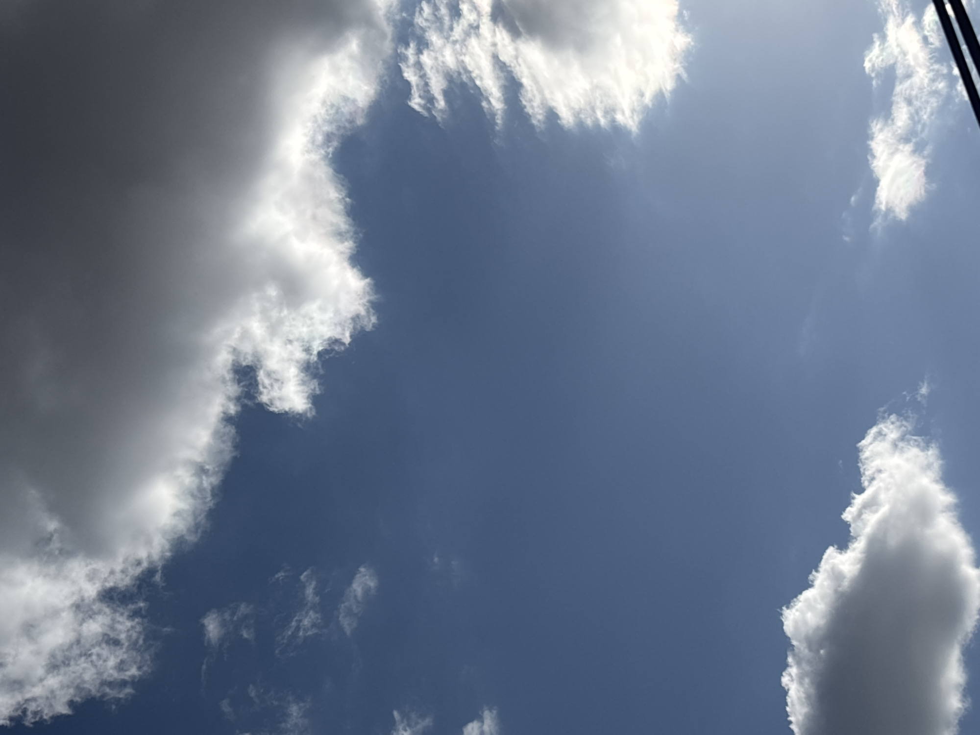 The image shows a view of the sky in Yerevan, Armenia. Predominantly blue, the sky is partially covered by white and grey clouds. A dark object, possibly power lines or tree branches, is visible in the upper right corner.