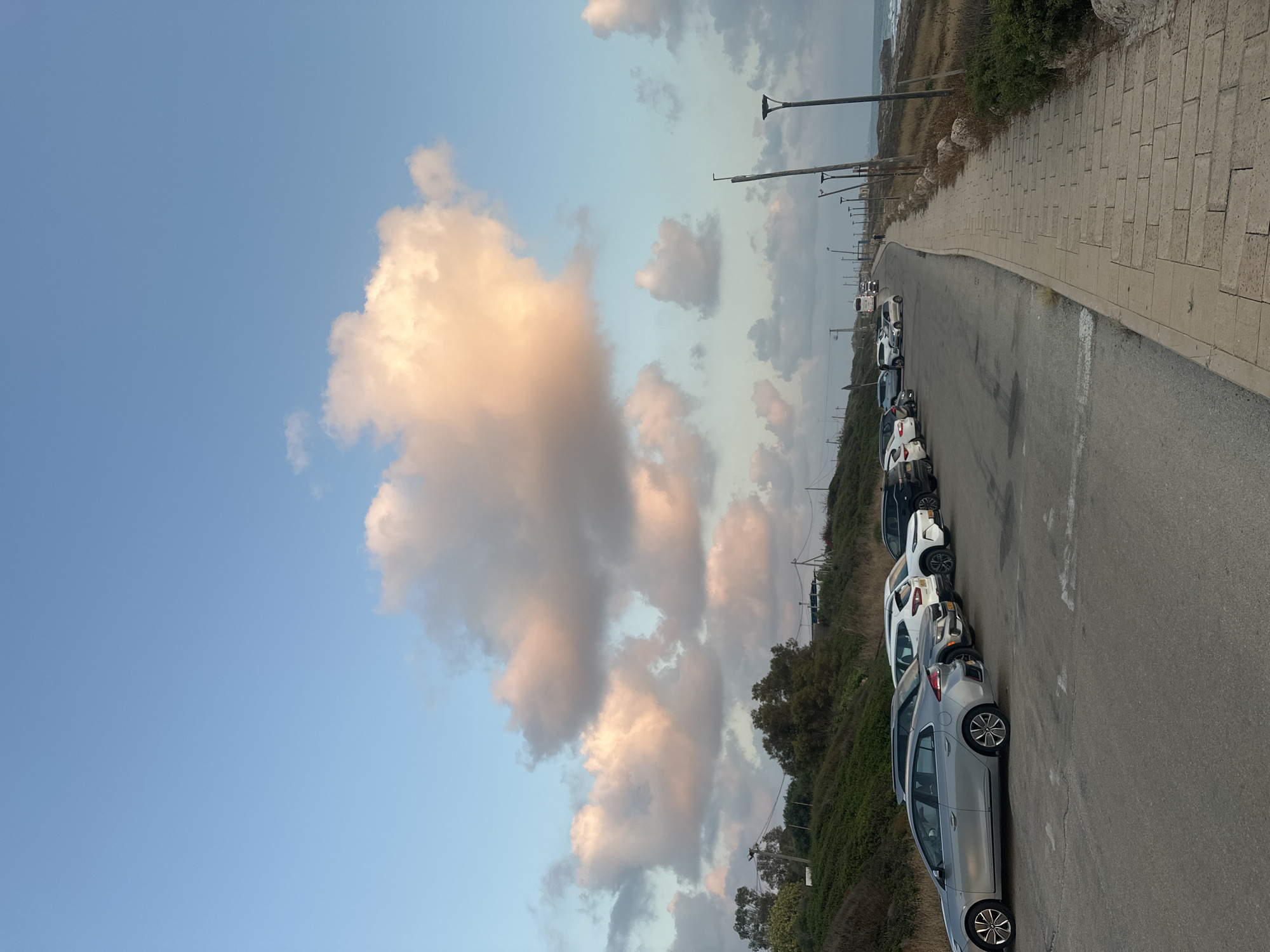 The image captures a scene in the Mate Asher Regional Council, Israel, likely during sunset, given the warm hues of the clouds. In the foreground, a paved road curves to the right, bordered by a stone sidewalk on the right and a grassy, slightly elevated embankment on the left. A line of parked cars, including a silver sedan and several SUVs and compact cars, are visible on the left side of the road. Beyond the cars, the embankment is covered in green vegetation and scattered trees. Further down the road, several streetlights are positioned along the sidewalk. In the distance, a bus and other vehicles are traveling on the road. The sky is a prominent feature, filled with large, puffy clouds tinged with pink and orange, set against a clear blue sky. The sea is visible on the far right of the image, with waves lapping at the shore. The overall atmosphere is serene and natural.
